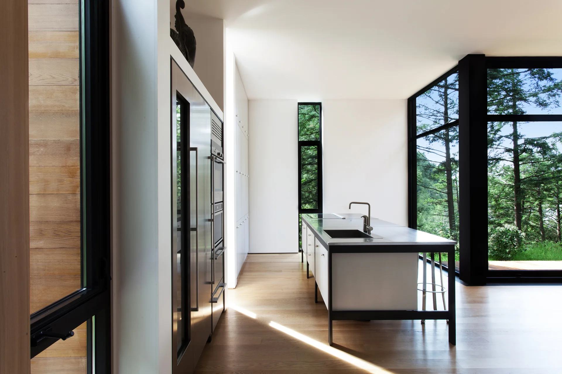 A minimalist kitchen with a stainless steel island, light wood flooring, and large black-framed windows displaying forest views.