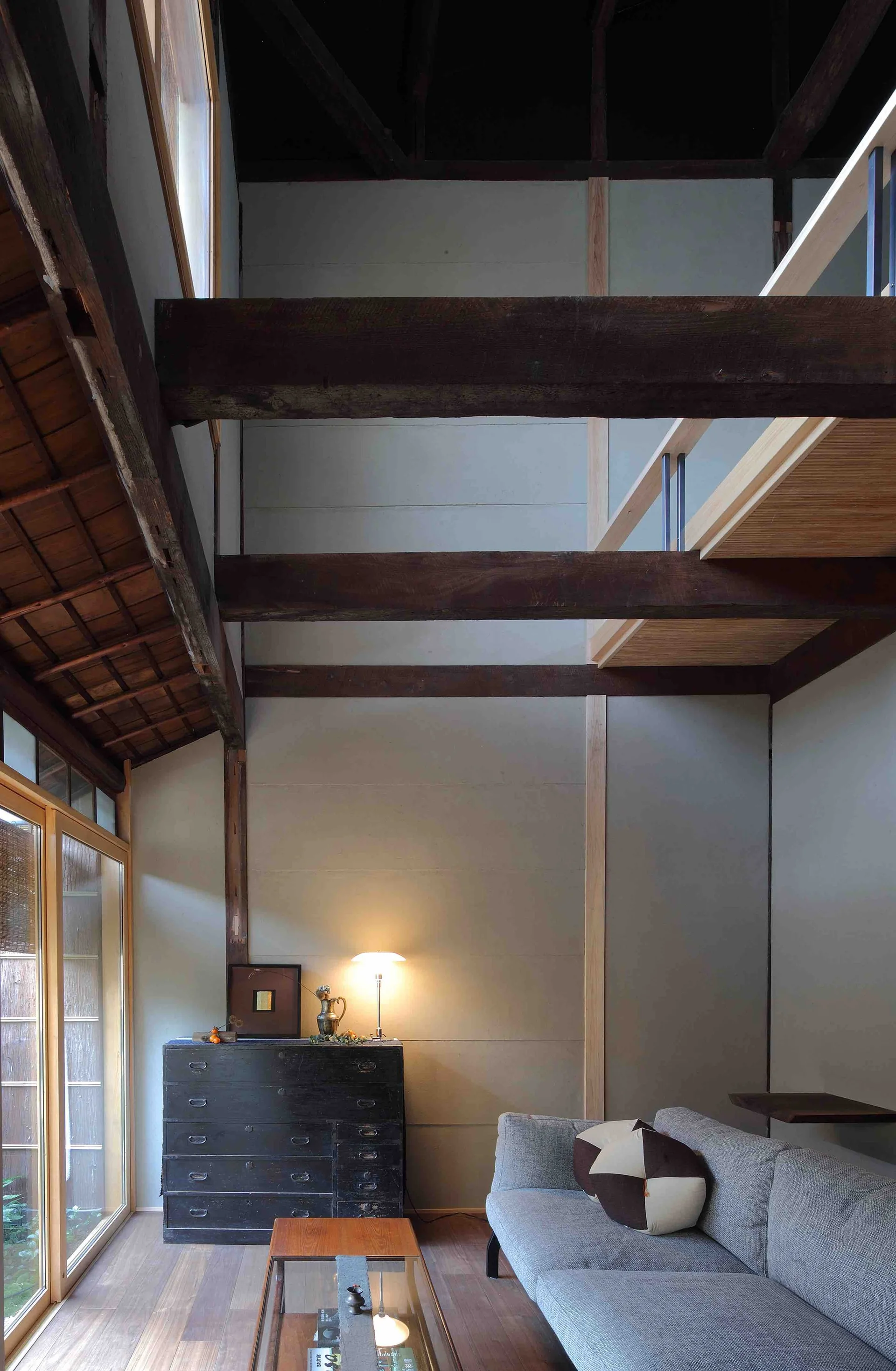 A rustic Japandi living room featuring a grey modern sofa, a dark antique chest of drawers, and high ceilings with exposed timber beams.