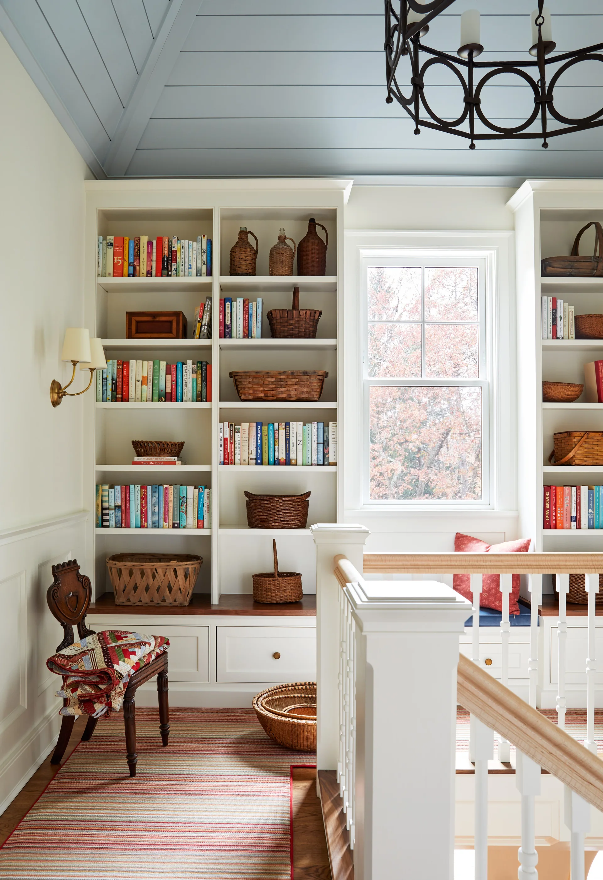 A bright staircase landing features white built-in bookshelves flanking a central window under a light blue wood-planked ceiling.