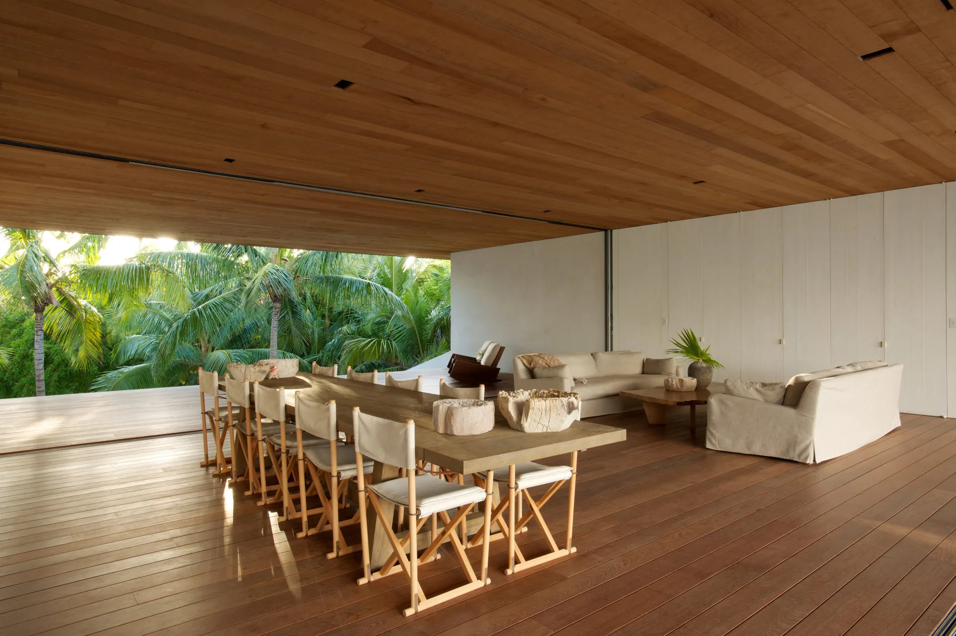 An open-concept indoor-outdoor living and dining area featuring a wood-plank ceiling, wooden dining table with canvas director's chairs, and beige sofas.