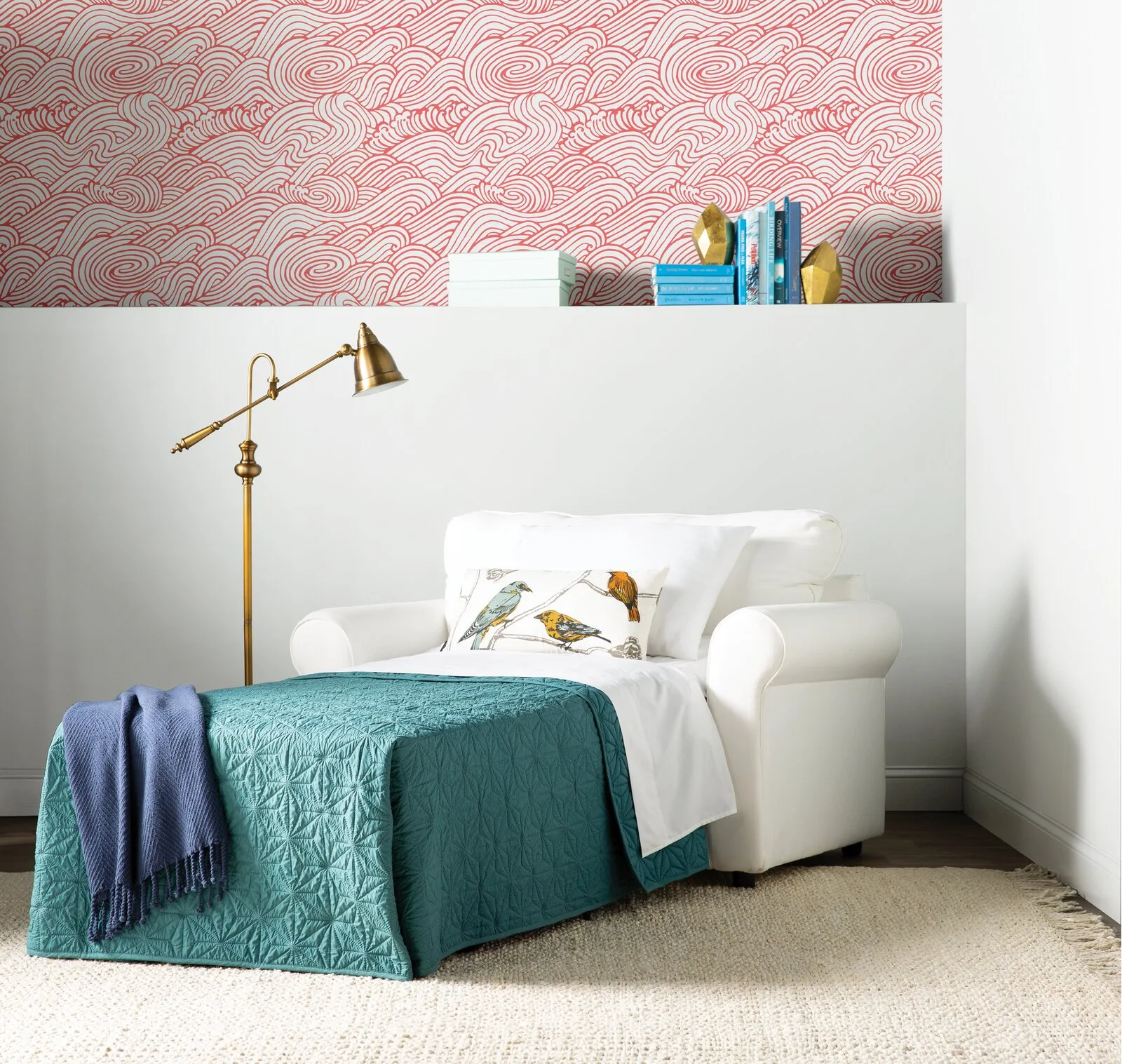 A white convertible sleeper chair is styled with a teal blanket next to a brass floor lamp under a red and white wave-patterned wallpapered wall.