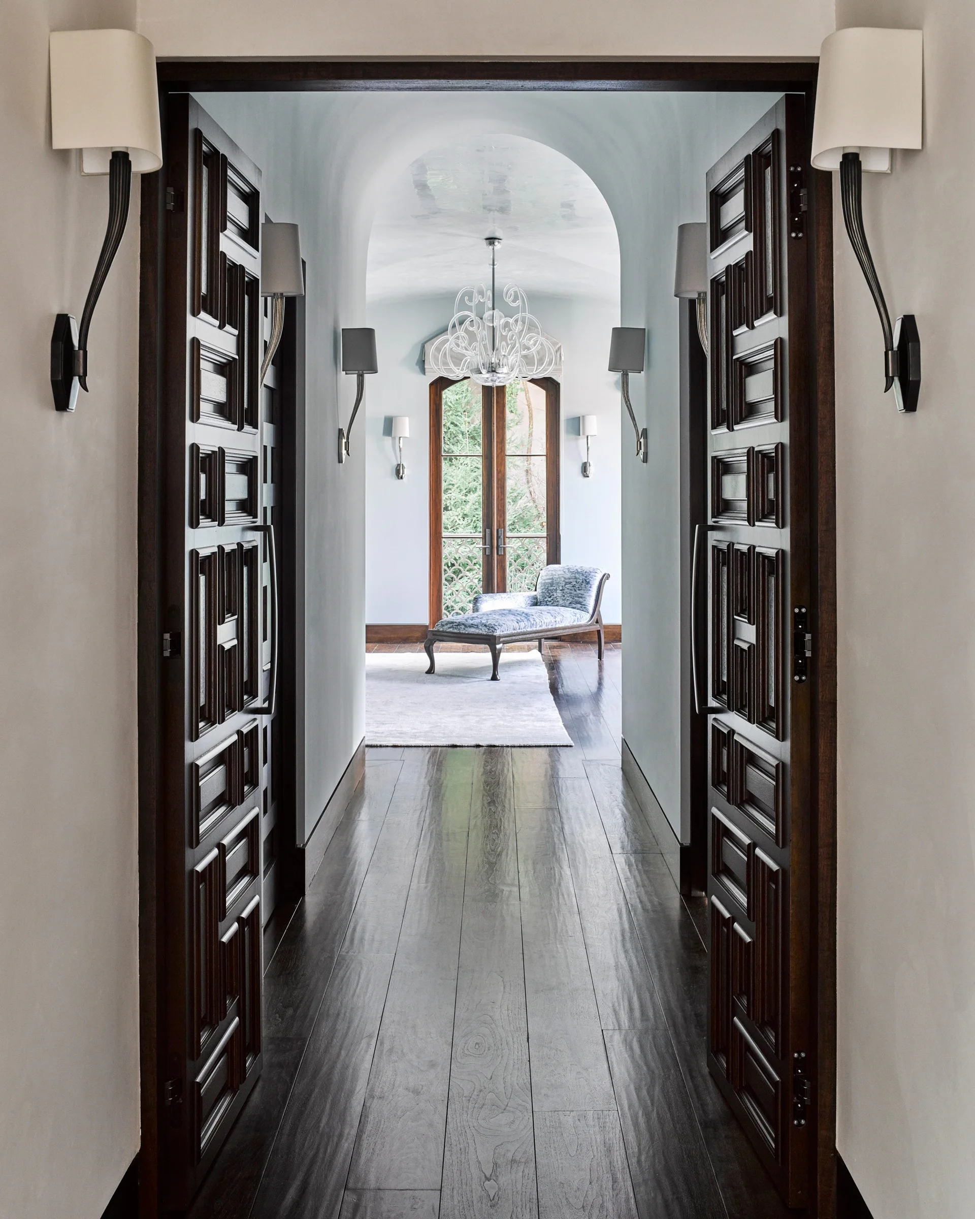 A view through geometric dark wood doors into an arched light blue hallway featuring wall sconces and leading to a chaise longue.