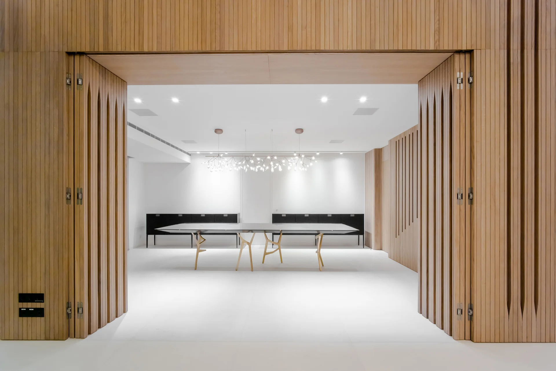 A minimalist dining room with a long table and branch chandelier is framed by textured vertical wood slat panels.