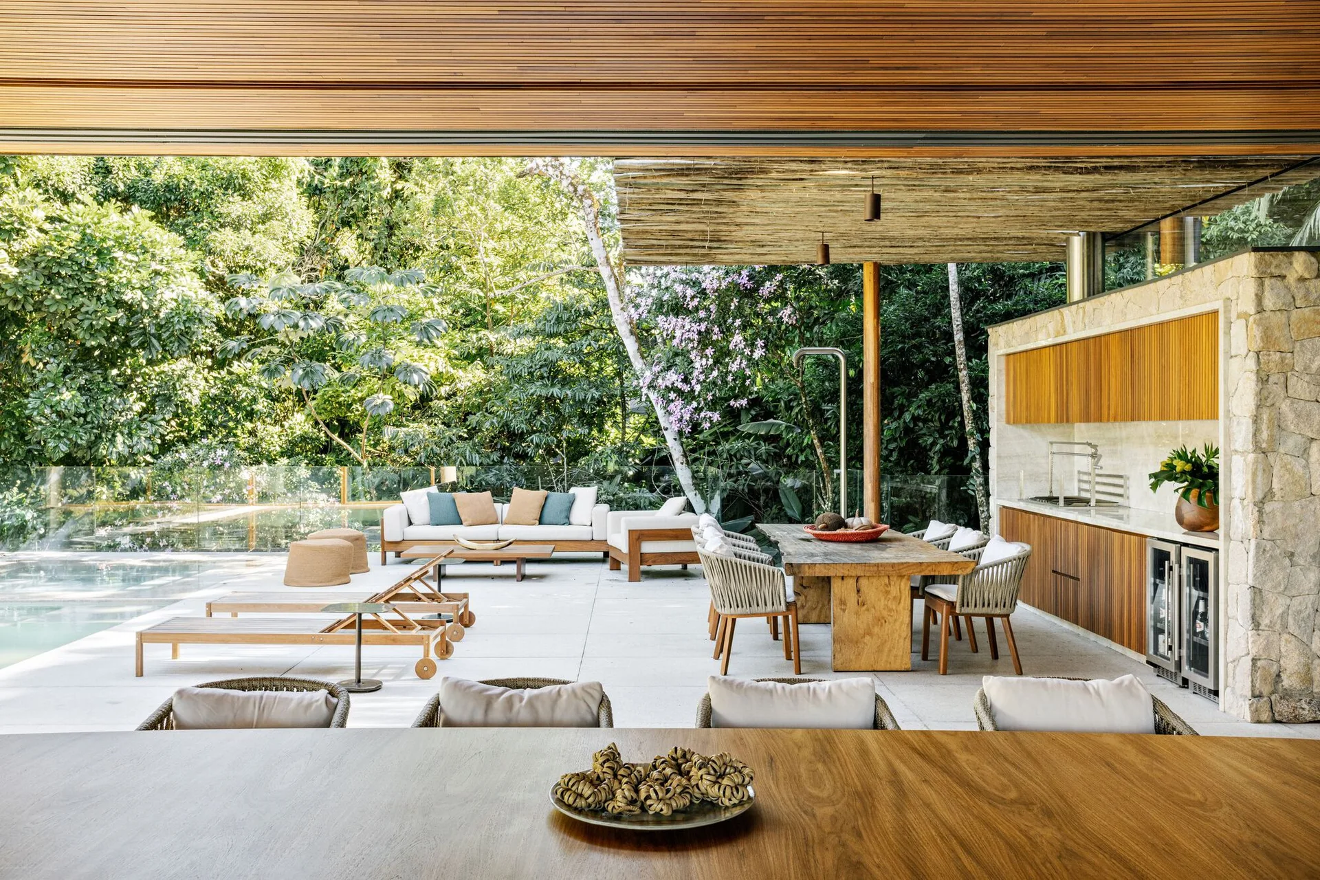 A covered outdoor patio features a slatted wood ceiling, a wooden dining set, a stone-clad outdoor kitchen, lounge seating, and a swimming pool.