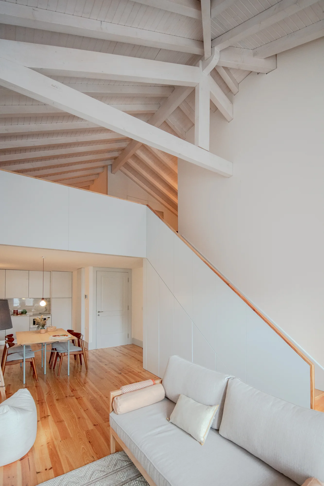 An open-plan living room features a white-painted vaulted ceiling with exposed wood beams, a light gray sofa, and a minimalist white staircase.