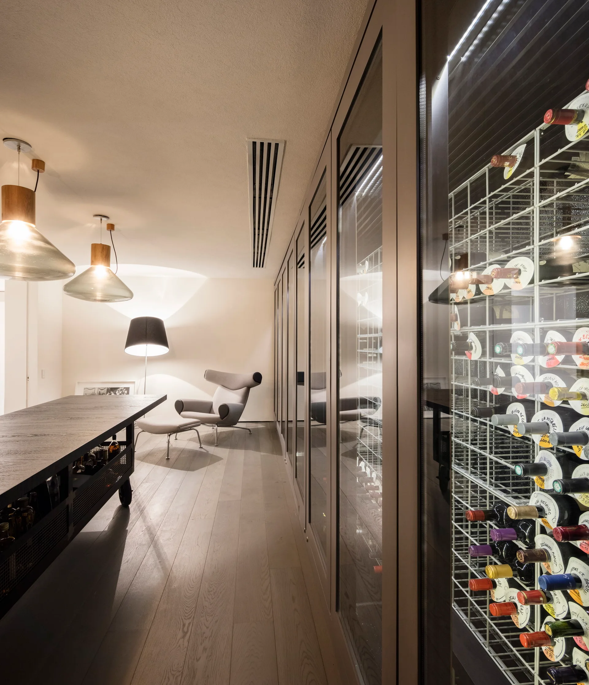 A modern wine room features a floor-to-ceiling glass-enclosed wine cabinet next to a lounge chair and a long dark wood table.