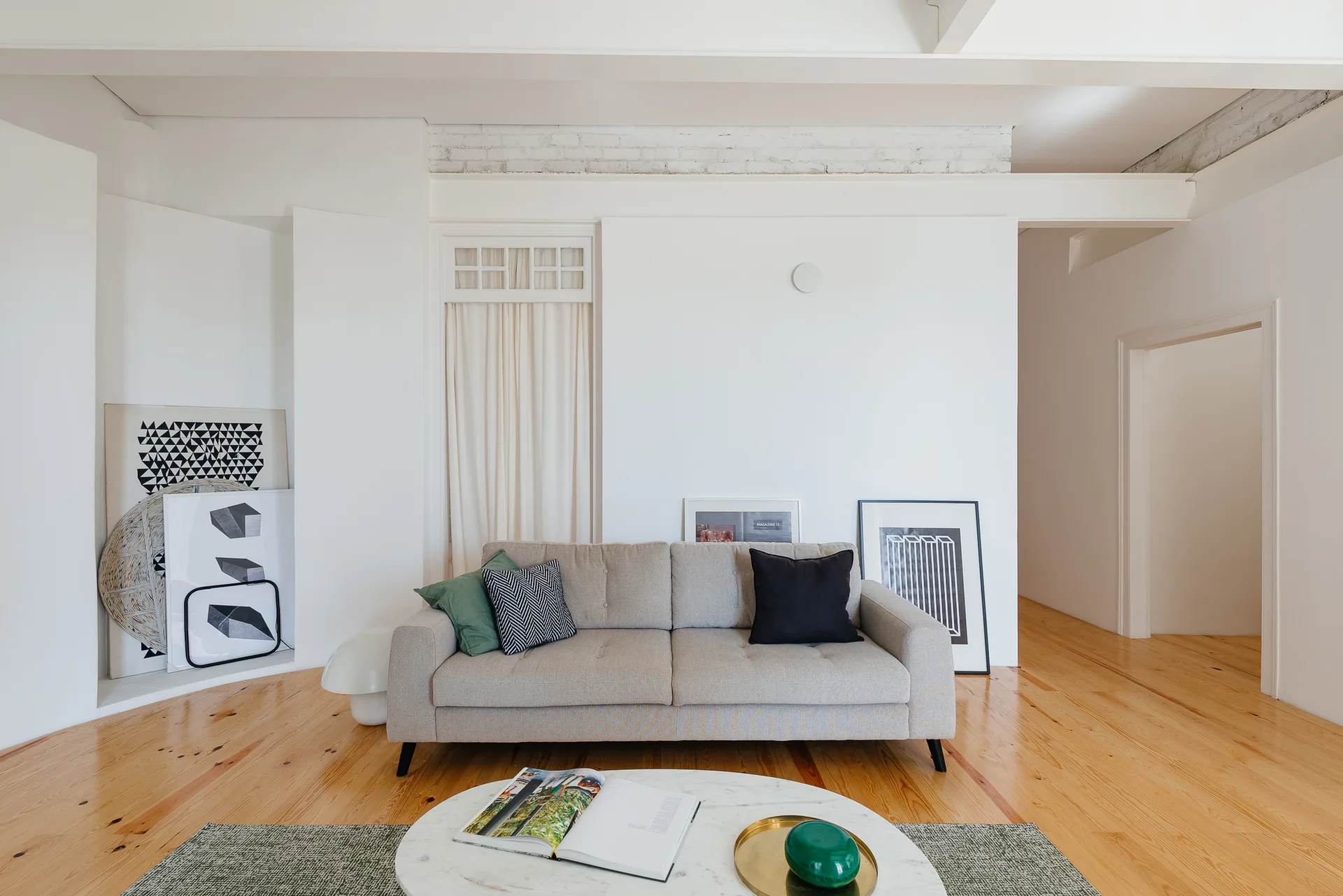 A light gray sofa sits on warm pine wood floors in a white living room with leaning graphic art and a round marble coffee table.