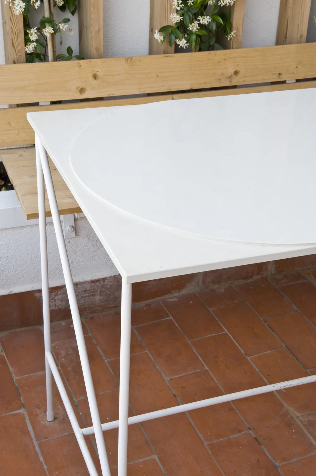 A white metal frame table with a circular top sits on a terracotta tiled patio next to a wooden slatted trellis.