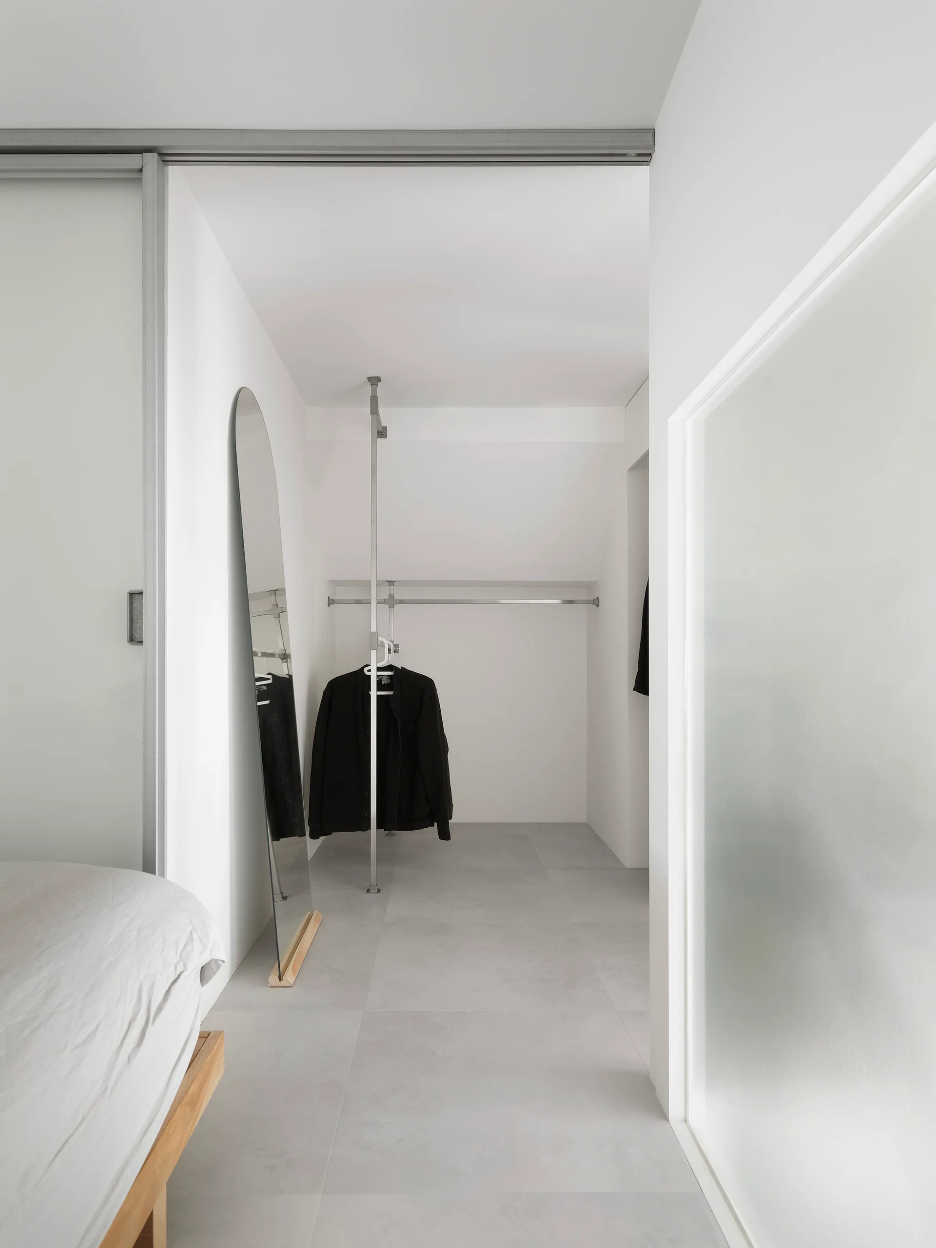 A minimalist walk-in closet seen from a bedroom, featuring a slim metal clothing rack, a tall arched floor mirror, and grey tile flooring.