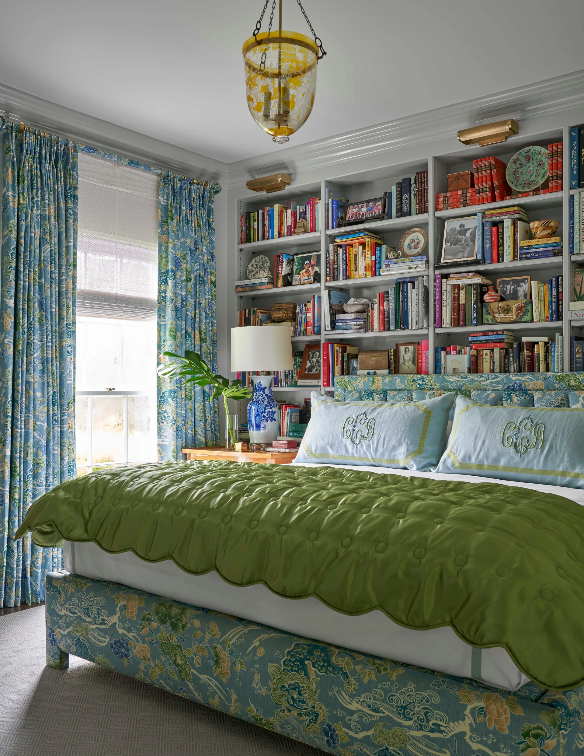A traditional bedroom featuring an upholstered blue floral bed with a green scalloped quilt set against a wall of built-in bookshelves.