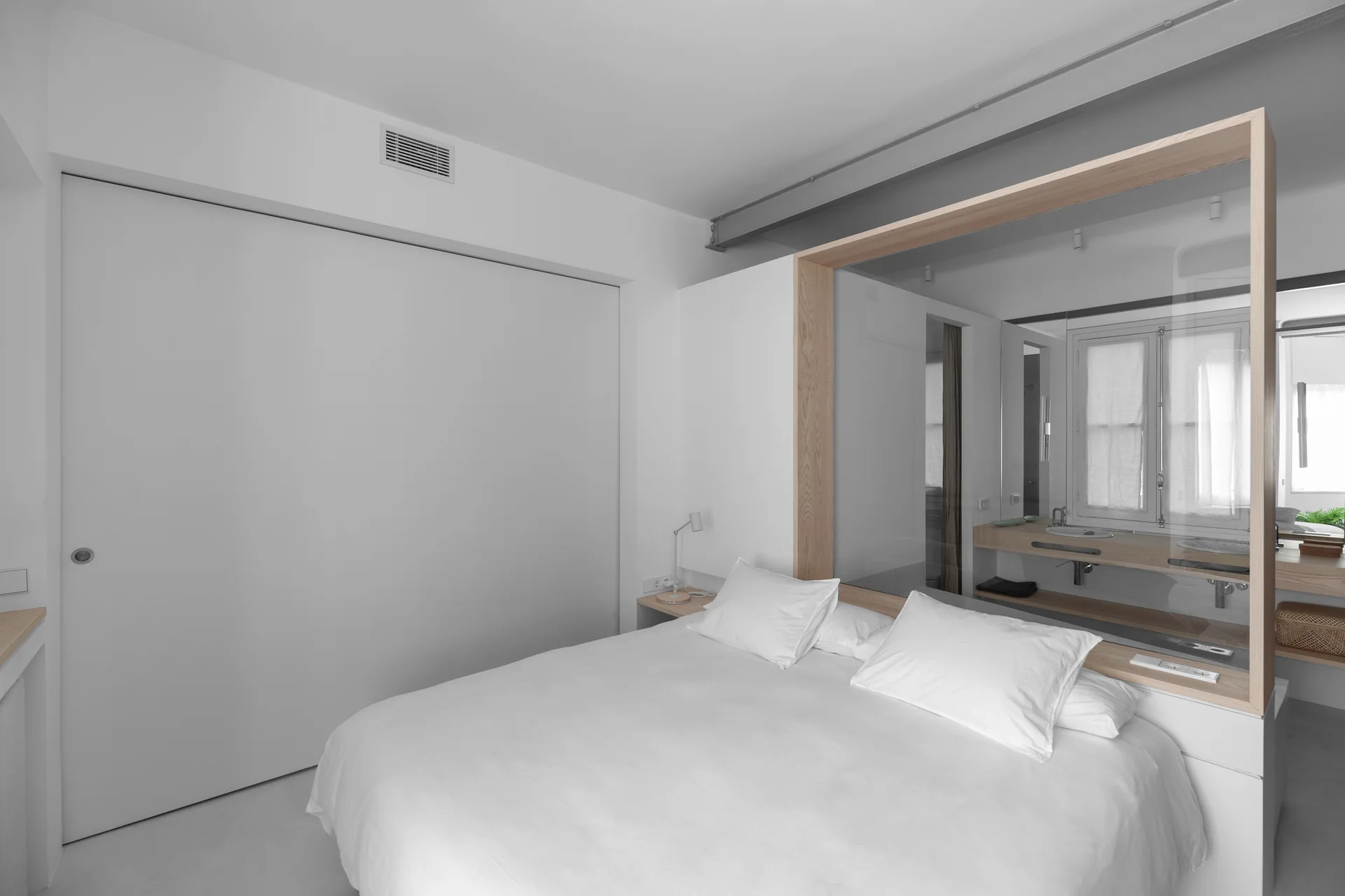 A minimalist white bedroom featuring a bed with white linens and a wood-framed glass partition separating the sleeping area from an ensuite bathroom.