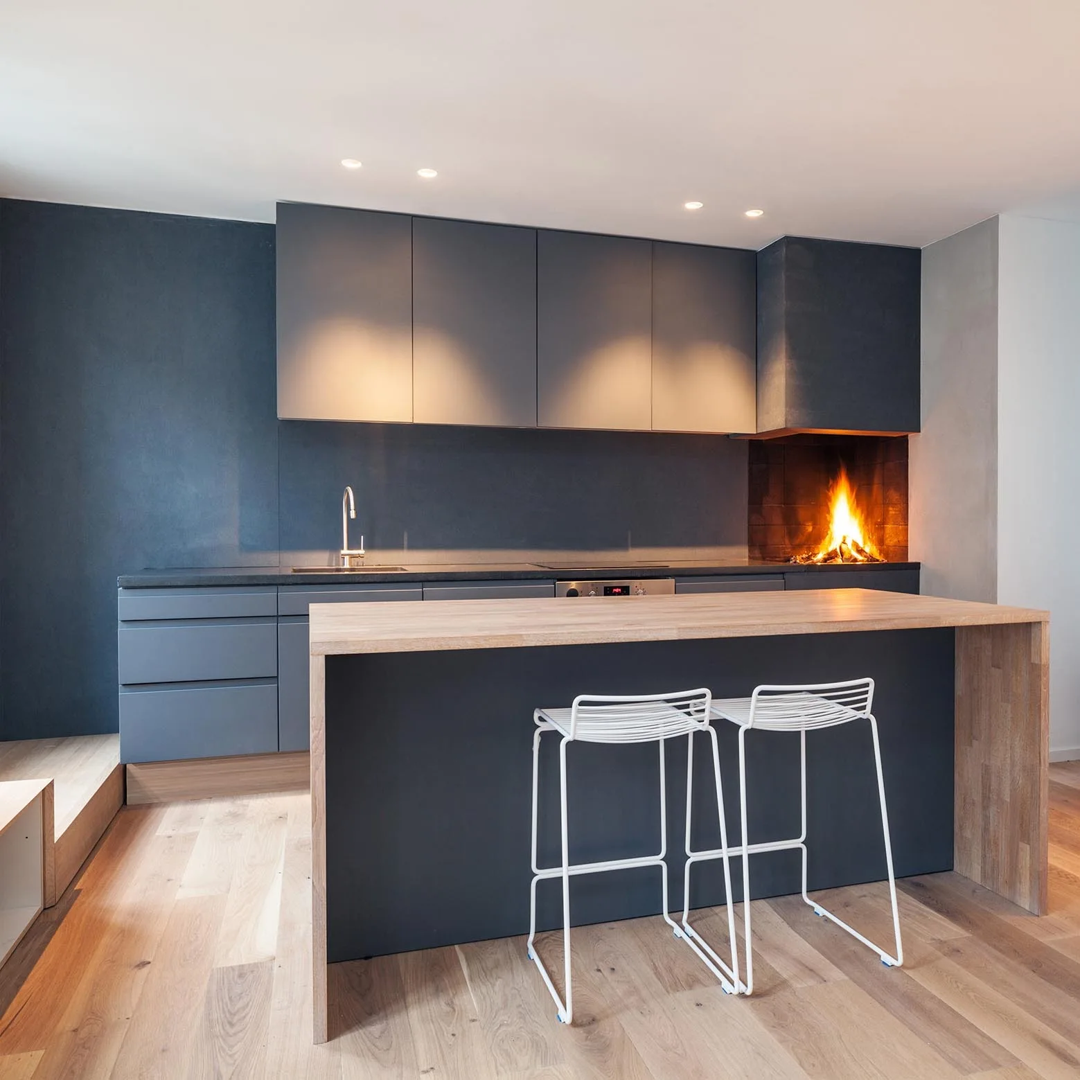 A modern kitchen features dark grey cabinets, a matching island with a wood-toned countertop, two white wire stools, and a built-in fireplace.