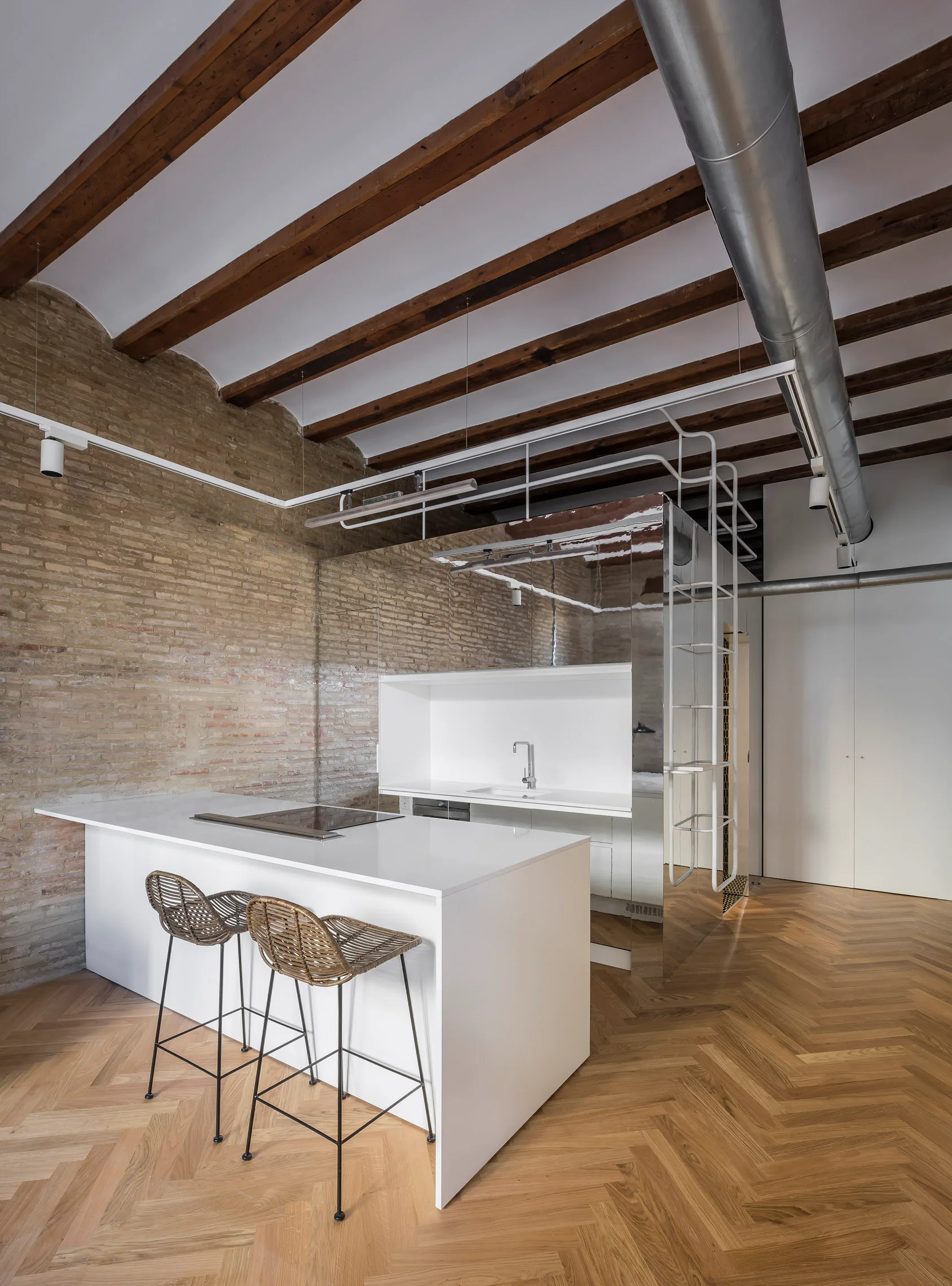 A minimalist kitchen features a white island with rattan barstools, a textured brick wall, herringbone wood floors, and exposed ceiling beams.