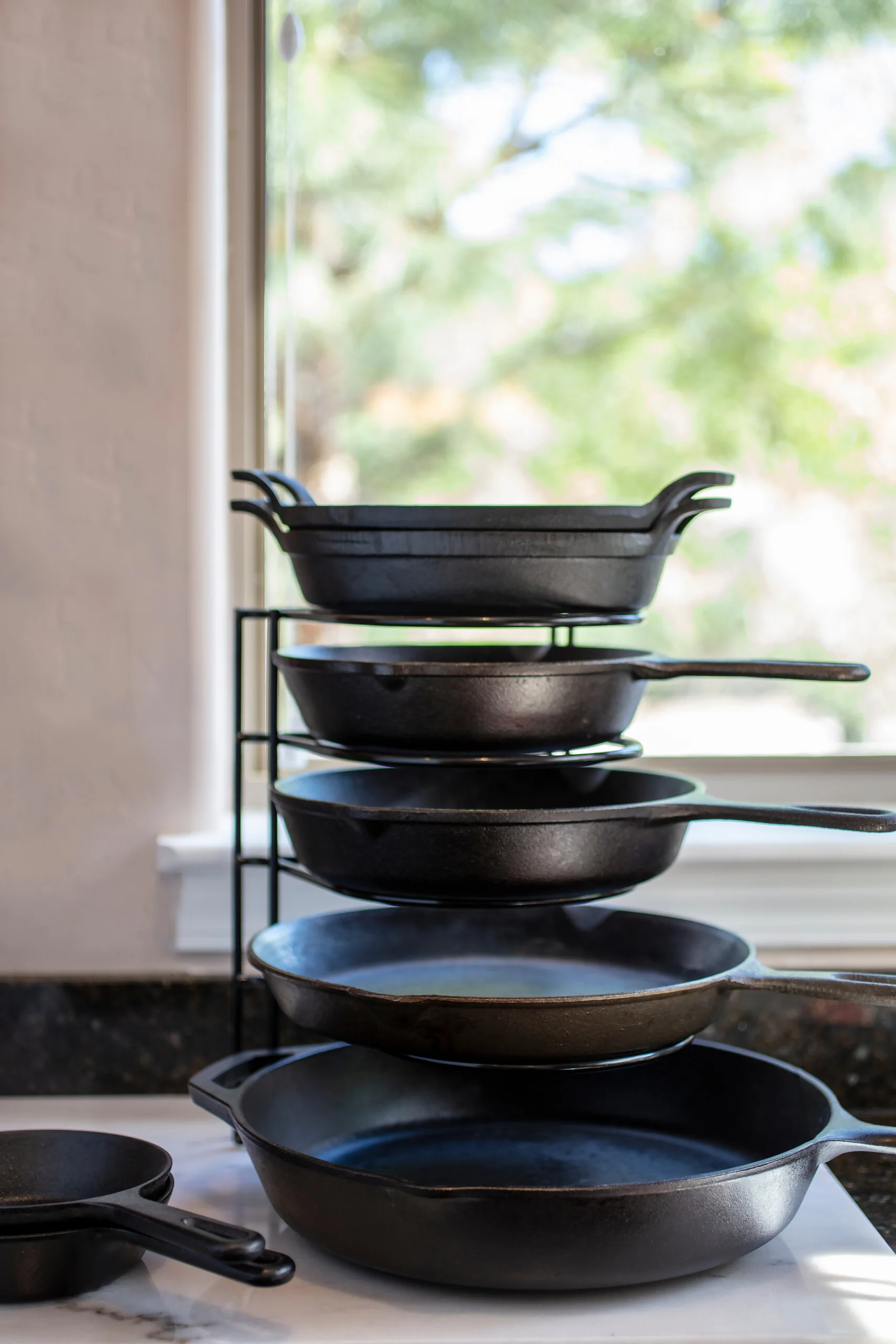 Multiple black cast iron skillets are stacked vertically on a black wire storage rack on a white marble countertop near a bright window.