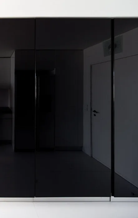 High-gloss black minimalist cabinet panels reflect a white room and door above a white floor.