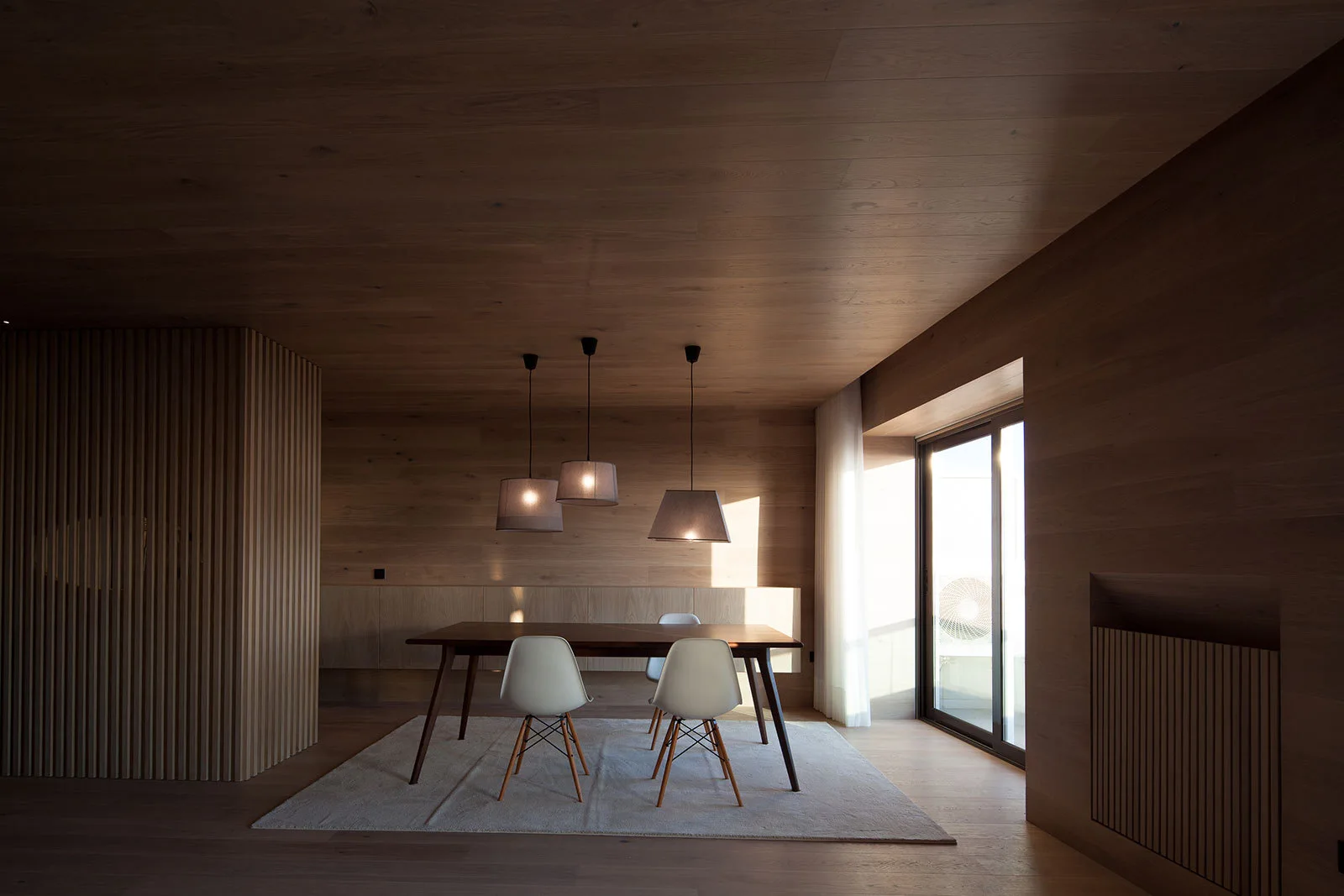 A minimalist dining room featuring wood-paneled walls and ceiling, a dark wood dining table, white chairs, and three pendant lights.