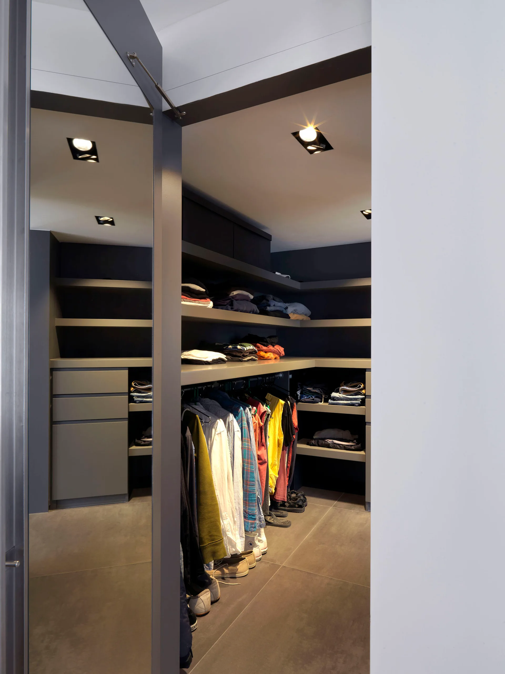 A modern walk-in closet with dark grey built-in shelves, hanging clothes, recessed ceiling lights, and a full-length mirrored door.