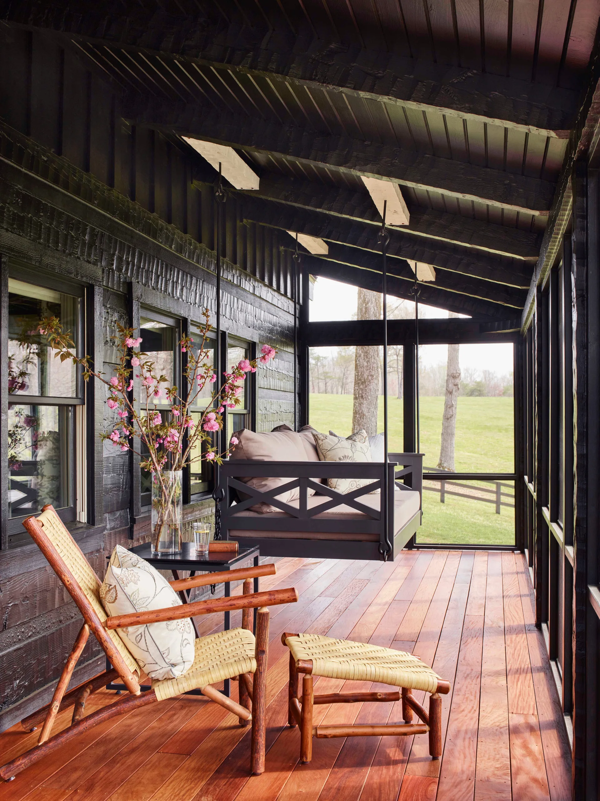 A screened porch features a black hanging daybed swing, vertical dark siding, a rustic wood lounge chair, and warm plank flooring.