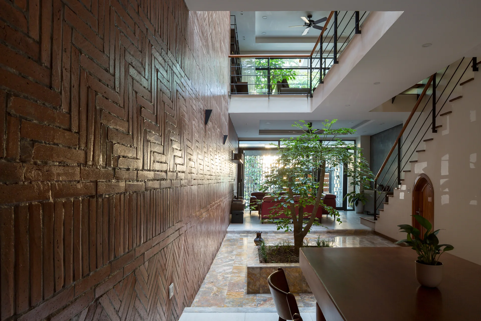 A multi-level home interior featuring a long patterned red brick wall, an indoor courtyard with a tree, stone tile floors, and a wooden staircase.
