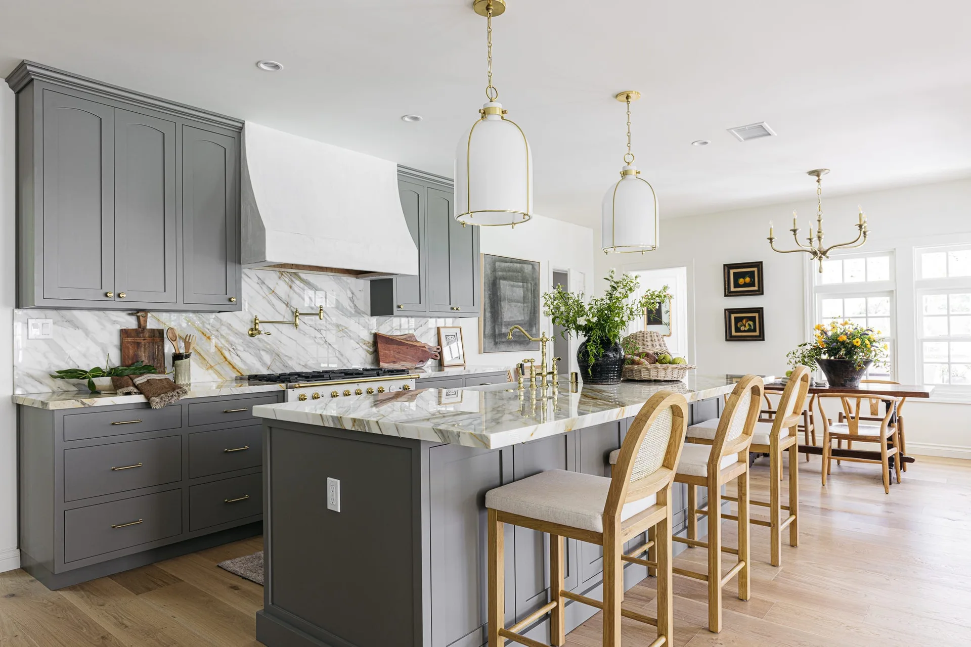 A transitional kitchen features grey cabinets, a white plaster range hood, a marble-topped island with cane bar stools, and brass pendant lights.