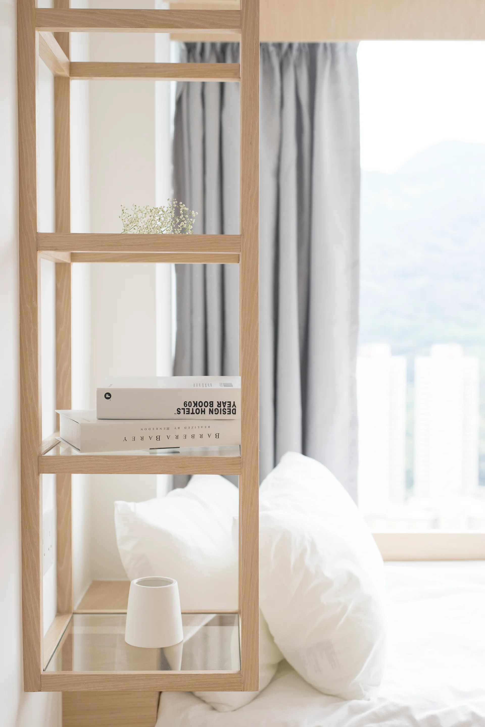 A light wood bedside shelving unit with glass shelves holds white books next to a bed with plush pillows and a grey curtain.