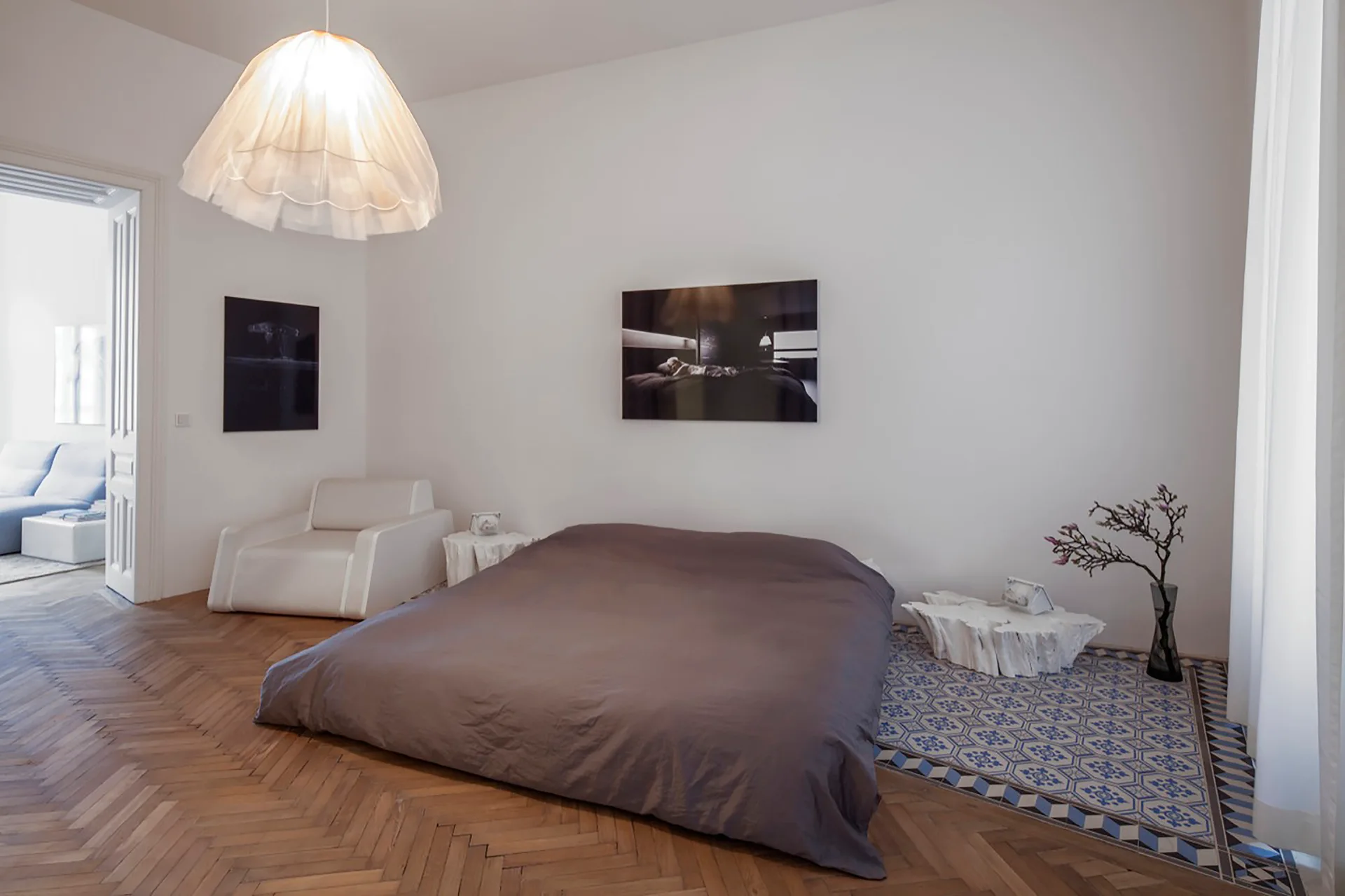 A low-profile bed sits in a minimalist white bedroom featuring herringbone hardwood floors and a blue geometric tile inlay.