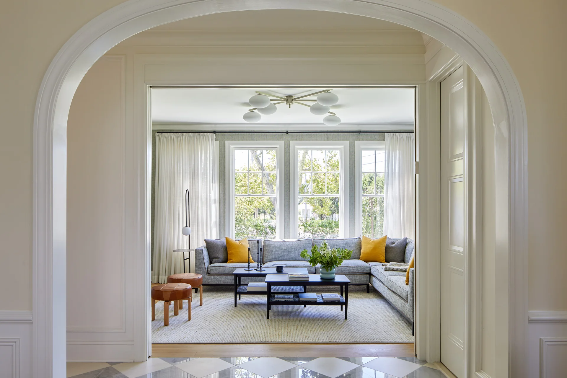 A light-filled living room with a large gray sectional sofa, yellow throw pillows, and two dark coffee tables, framed by a wide white plaster archway.