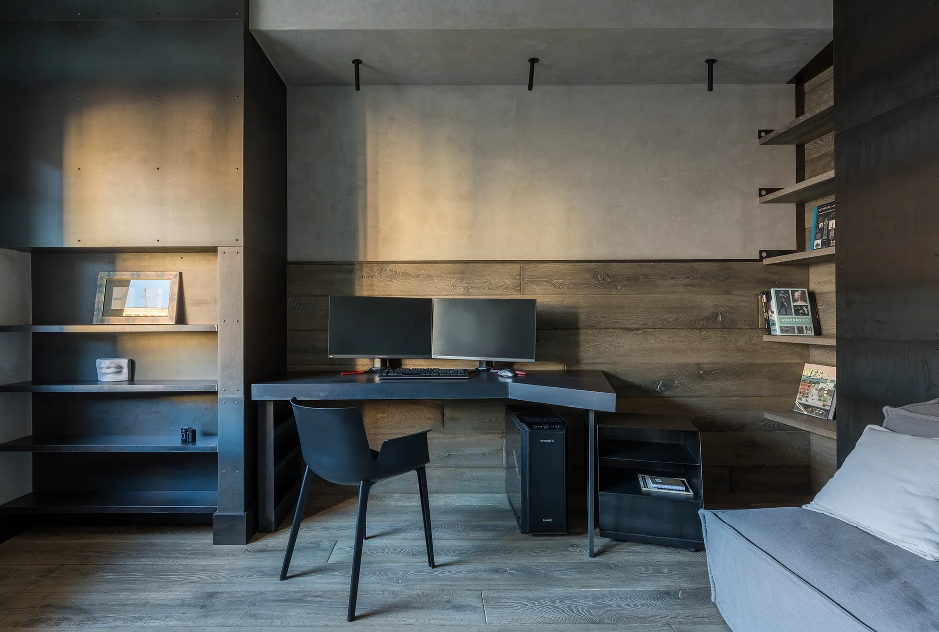 An industrial home office workstation with horizontal wood wall paneling, a dark angular metal desk, and custom built-in steel shelving.