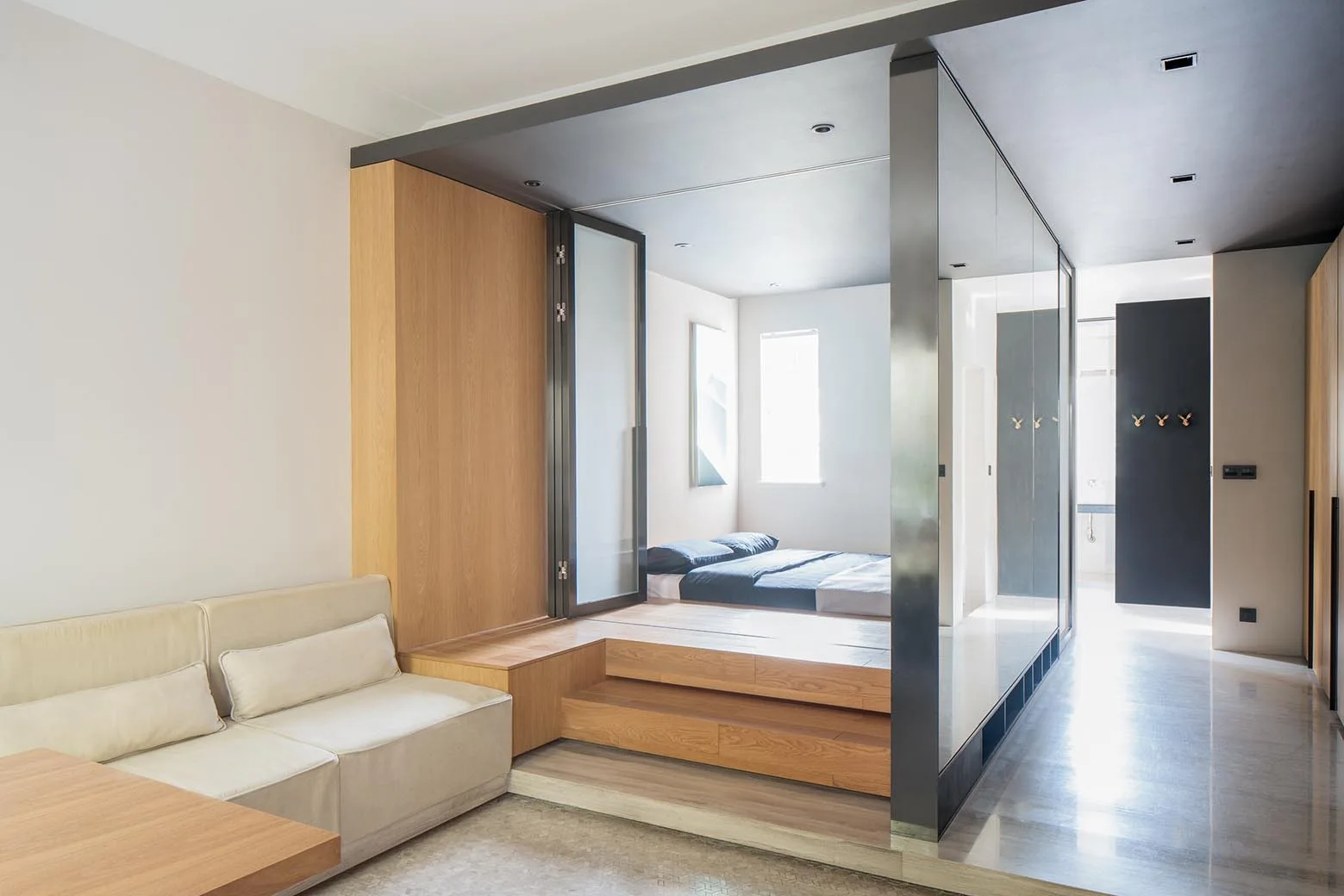 A modern open-plan studio featuring a raised wooden platform bedroom enclosed in glass partitions next to a beige sofa.