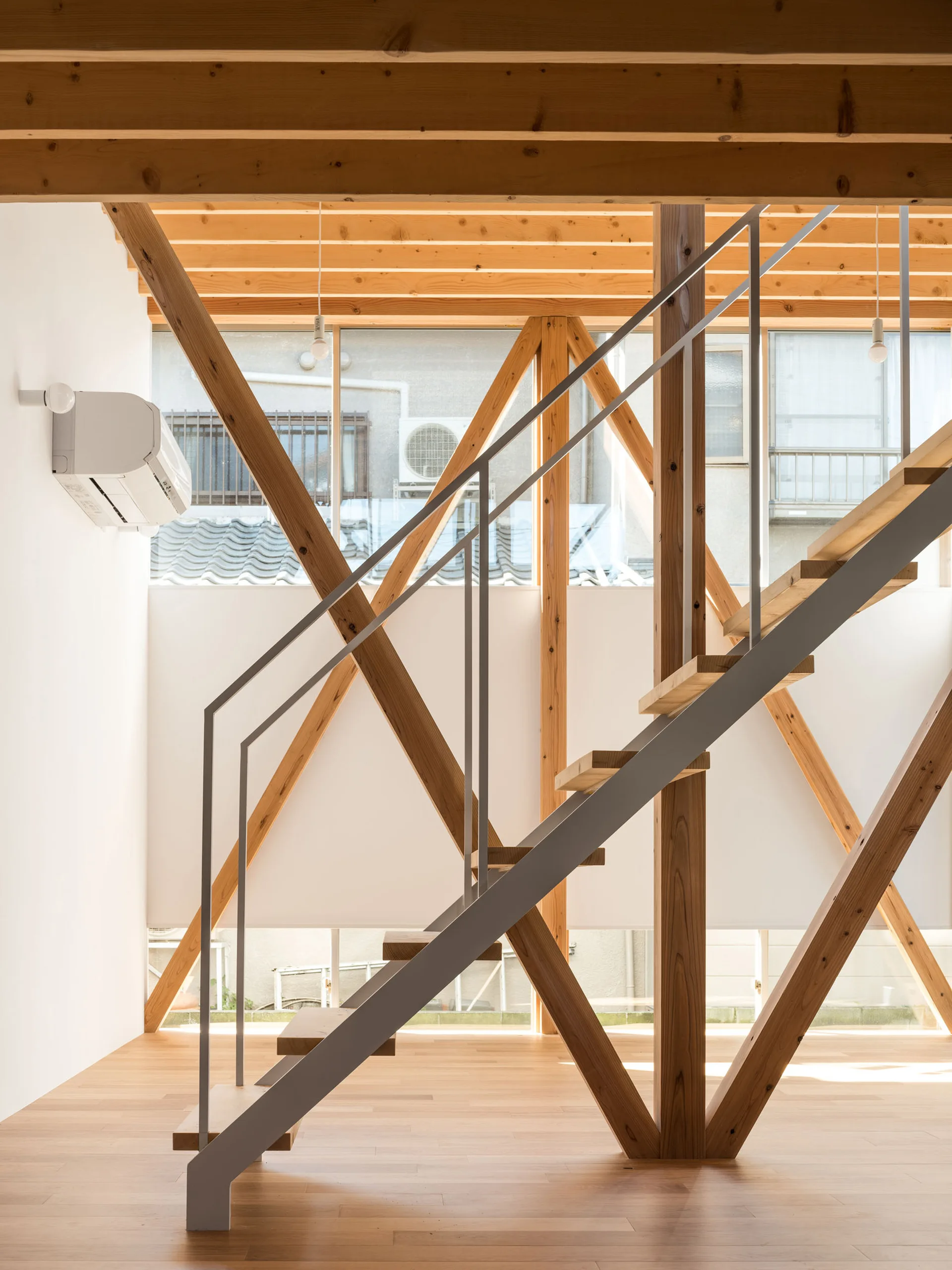 A minimalist interior featuring an open-riser wooden staircase with a grey metal railing, surrounded by exposed timber posts and beams.
