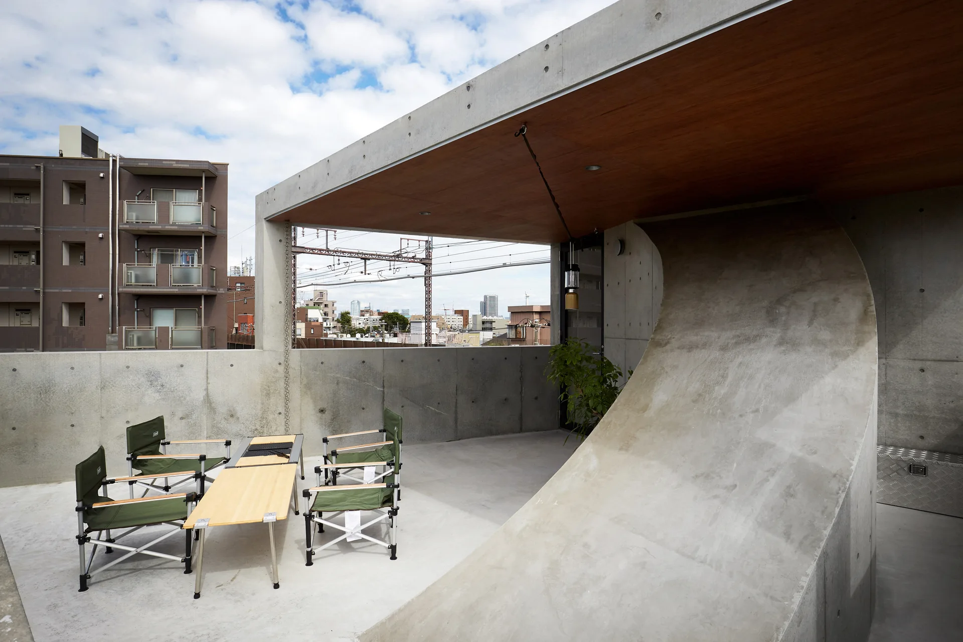 A concrete terrace features green folding chairs, a wood folding table, and a sculptural curved wall beneath a wood-paneled ceiling.