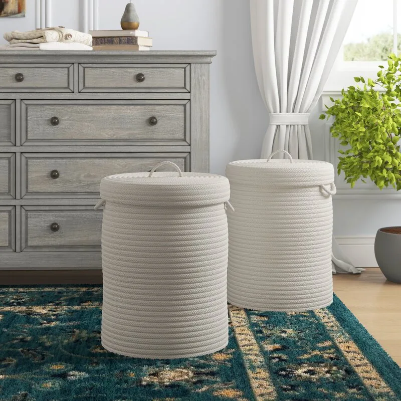 Two cylindrical cream-colored rope baskets with lids sit on a patterned teal area rug in front of a gray wooden dresser.