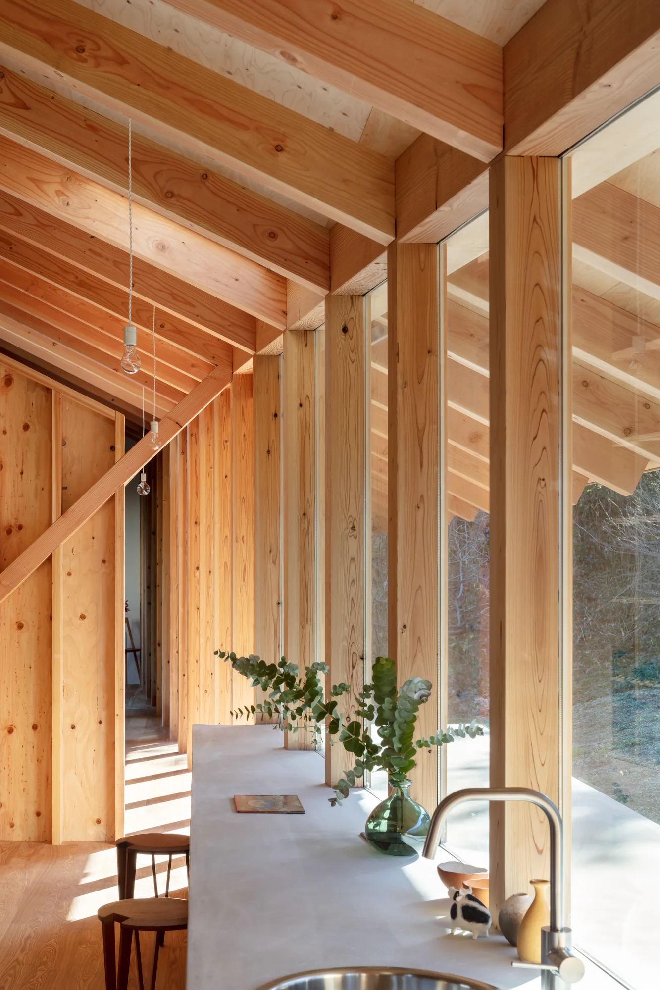 A minimalist kitchen corridor features a light grey countertop alongside large windows beneath exposed wooden ceiling rafters and vertical wood paneling.