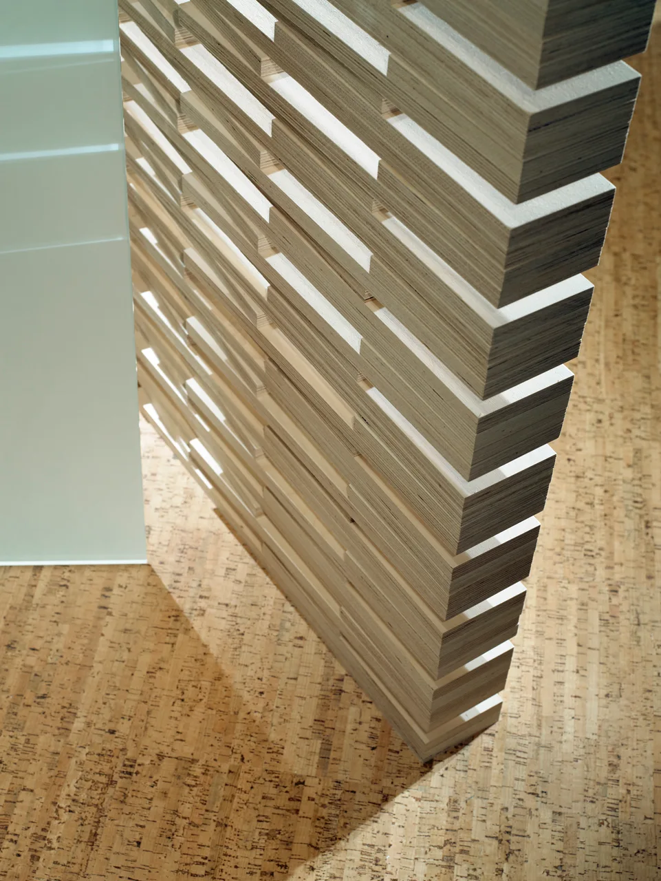 A close-up of a structured slatted plywood room divider standing on granular cork flooring next to a glass panel.