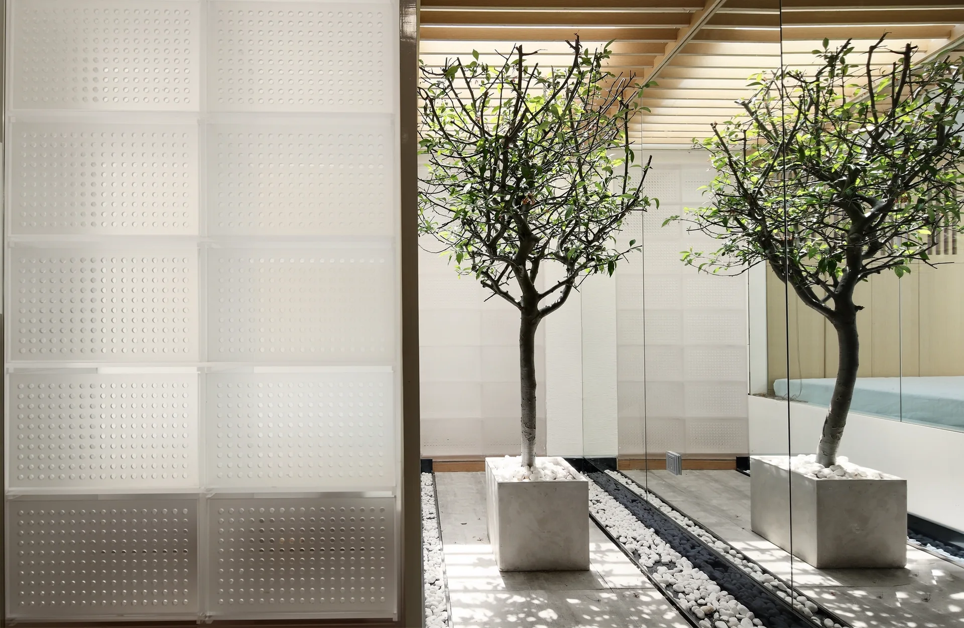 A minimalist indoor courtyard features a potted tree on a gravel bed next to a full-height mirror wall and a white perforated partition panel.
