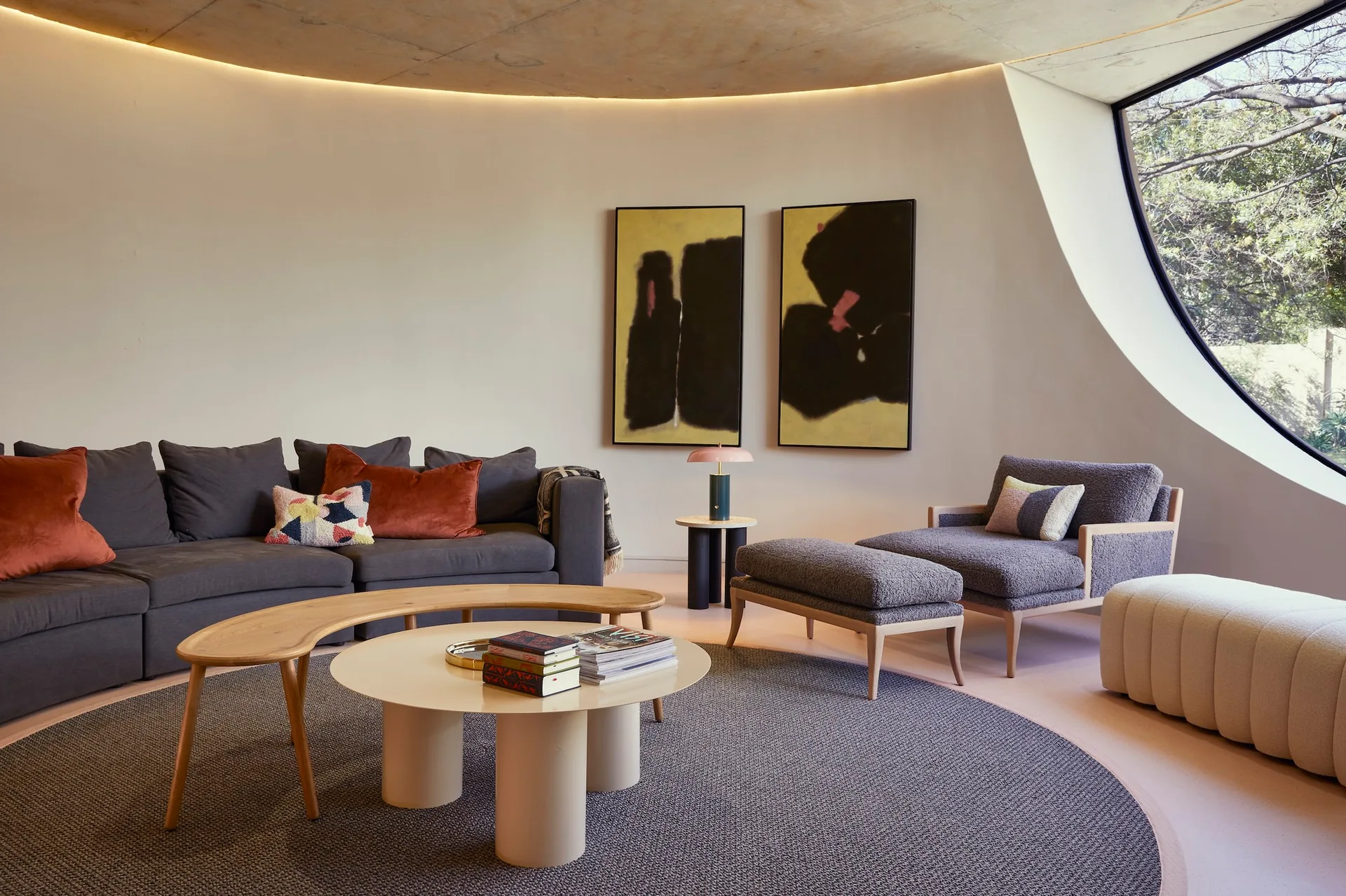 A contemporary living room features a curved off-white wall illuminated by cove lighting, a dark grey sectional, and a textured armchair with an ottoman.