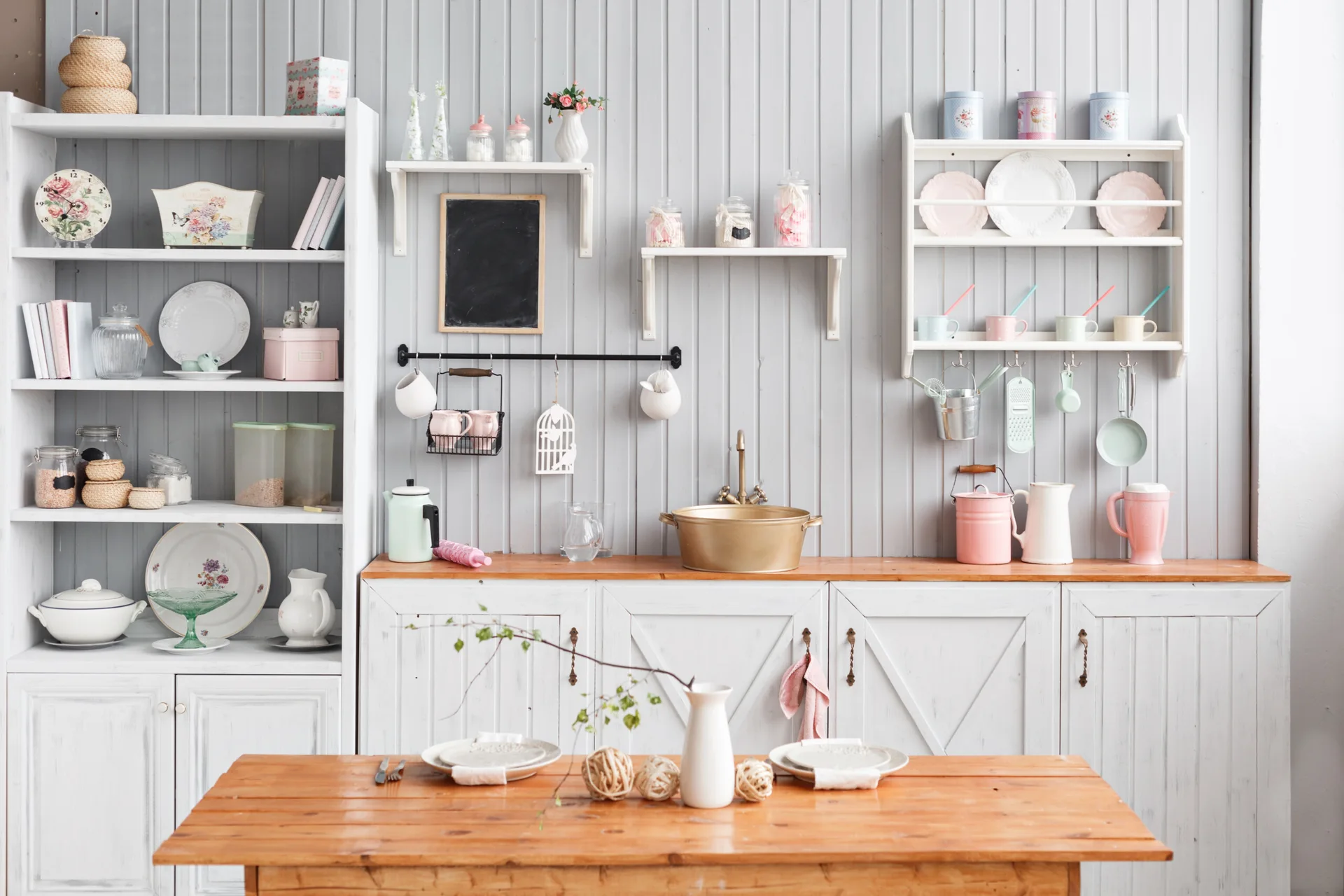 A cottage kitchen features grey vertical beadboard walls, white cabinets with a wood countertop, and a brass basin sink with open shelving.