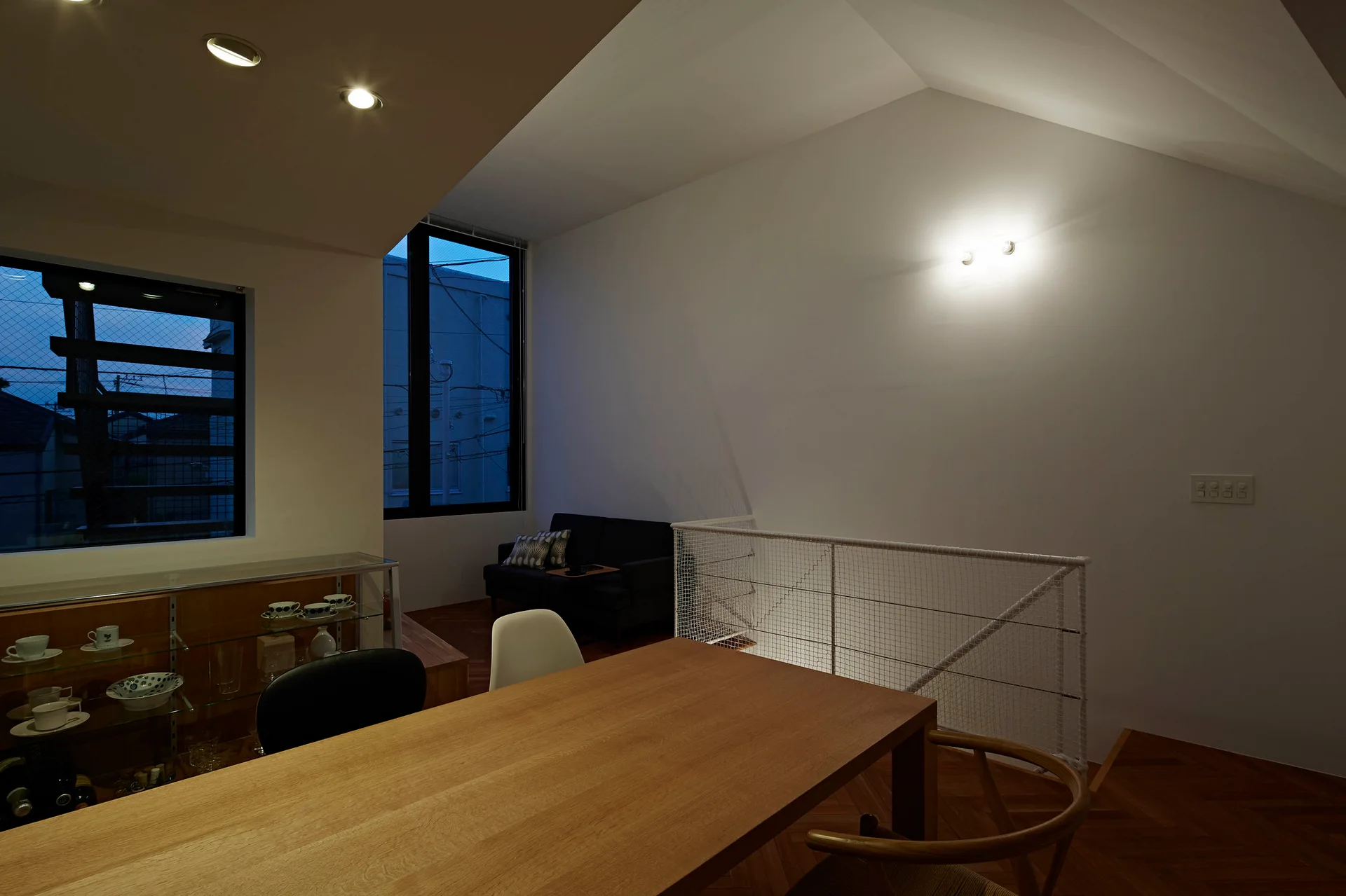 A minimalist dining room features a long wooden table, mismatched chairs, and a white mesh staircase railing beneath a high vaulted ceiling.