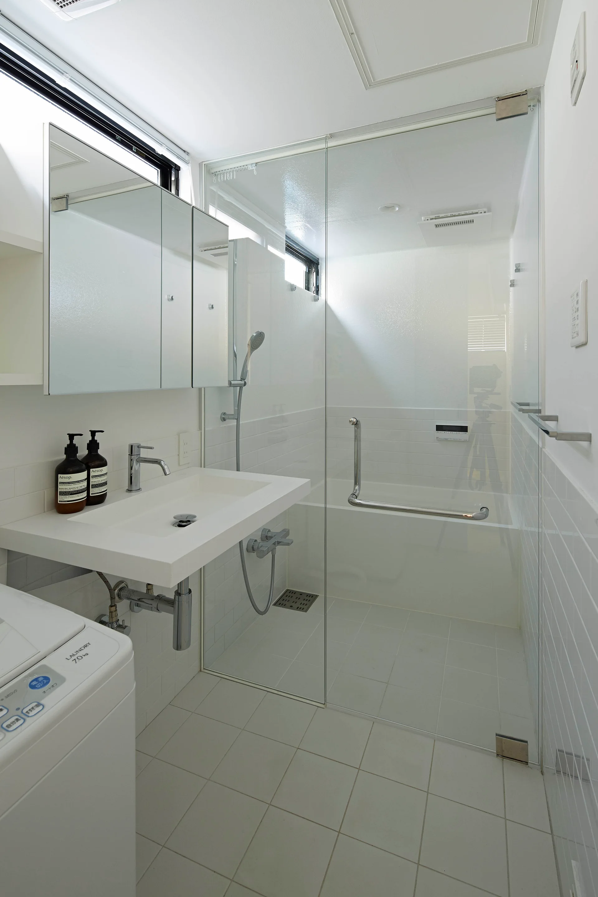 A minimalist white bathroom features a wall-mounted sink, mirrored medicine cabinets, and a glass-enclosed walk-in shower.