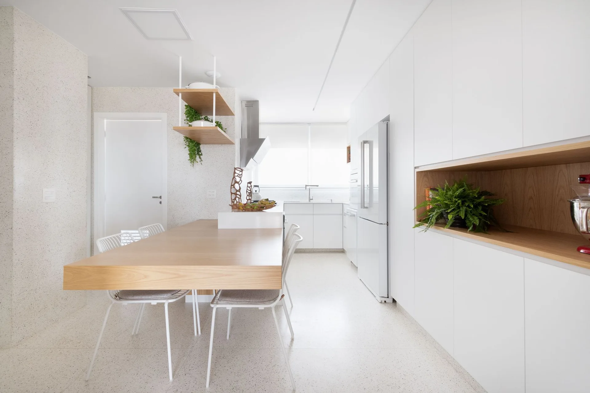 A bright minimalist kitchen features flat-front white cabinets, a light wood dining table integrated with the island, and speckled terrazzo flooring.