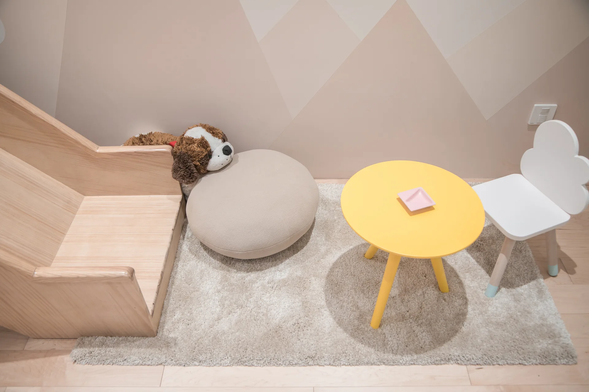 A kids play area features a small yellow table, a white cloud backrest chair, and a wooden floor seat on a soft beige rug.