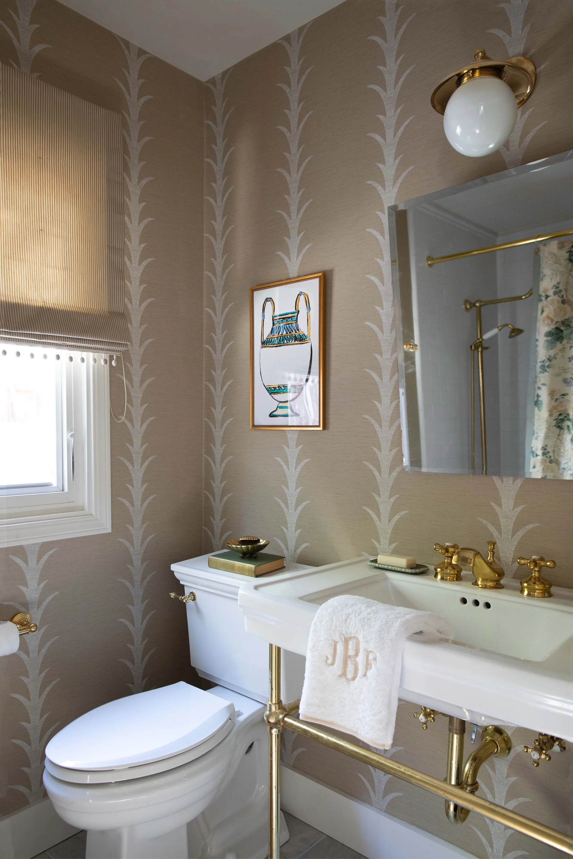 A powder room featuring beige patterned wallpaper, a white console sink with brass legs, and a toilet next to a window with a striped shade.