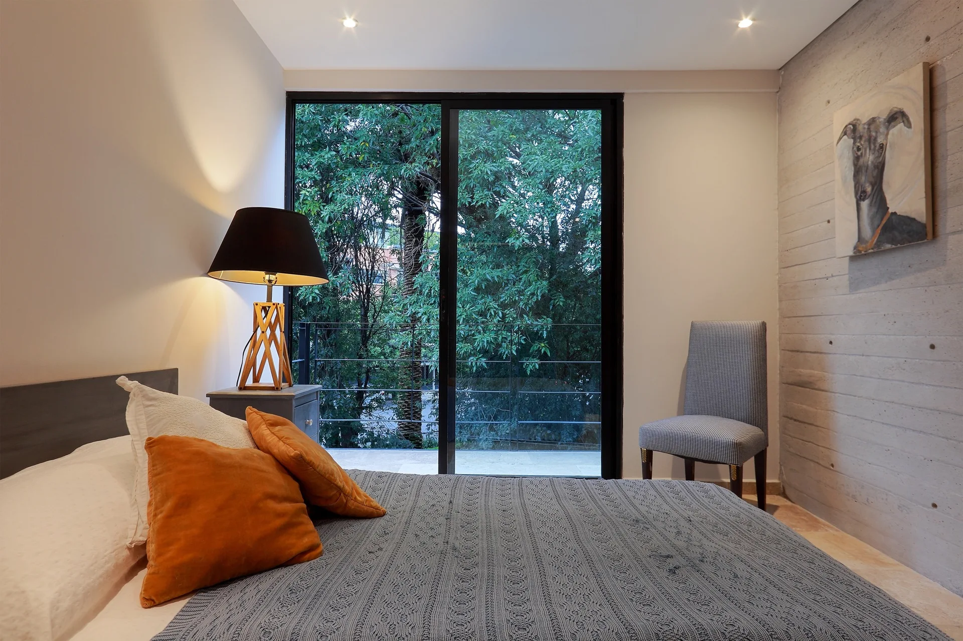 A modern bedroom features a textured concrete accent wall, a bed with orange throw pillows, and sliding glass doors looking out to lush green trees.