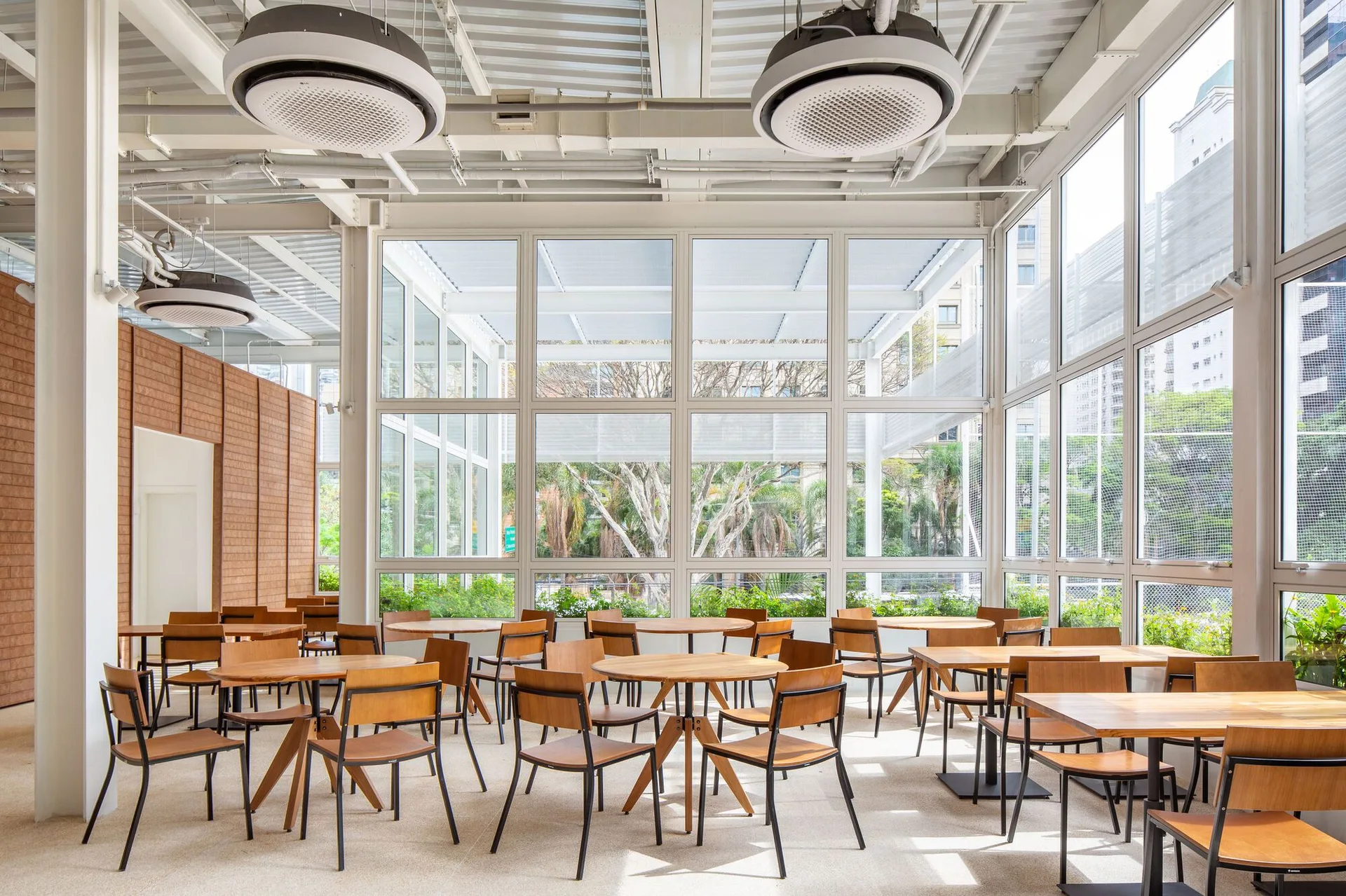 A bright commercial dining area with wooden tables and chairs set against large grid windows under an industrial exposed ceiling.