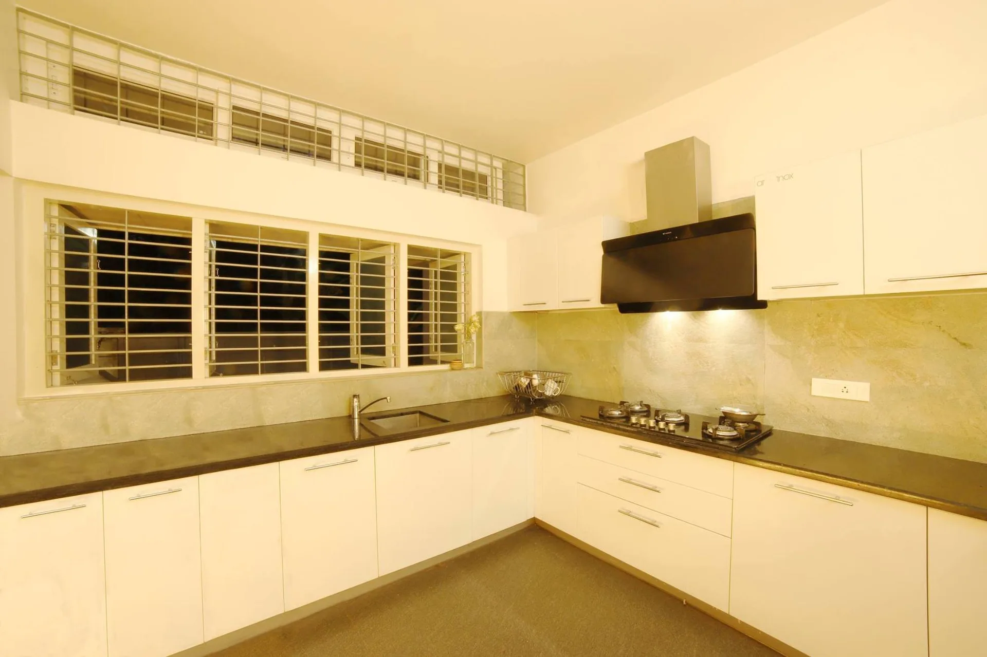 An L-shaped kitchen featuring flat-front white cabinets, dark countertops, a black range hood, and a large multi-pane window with security grates.