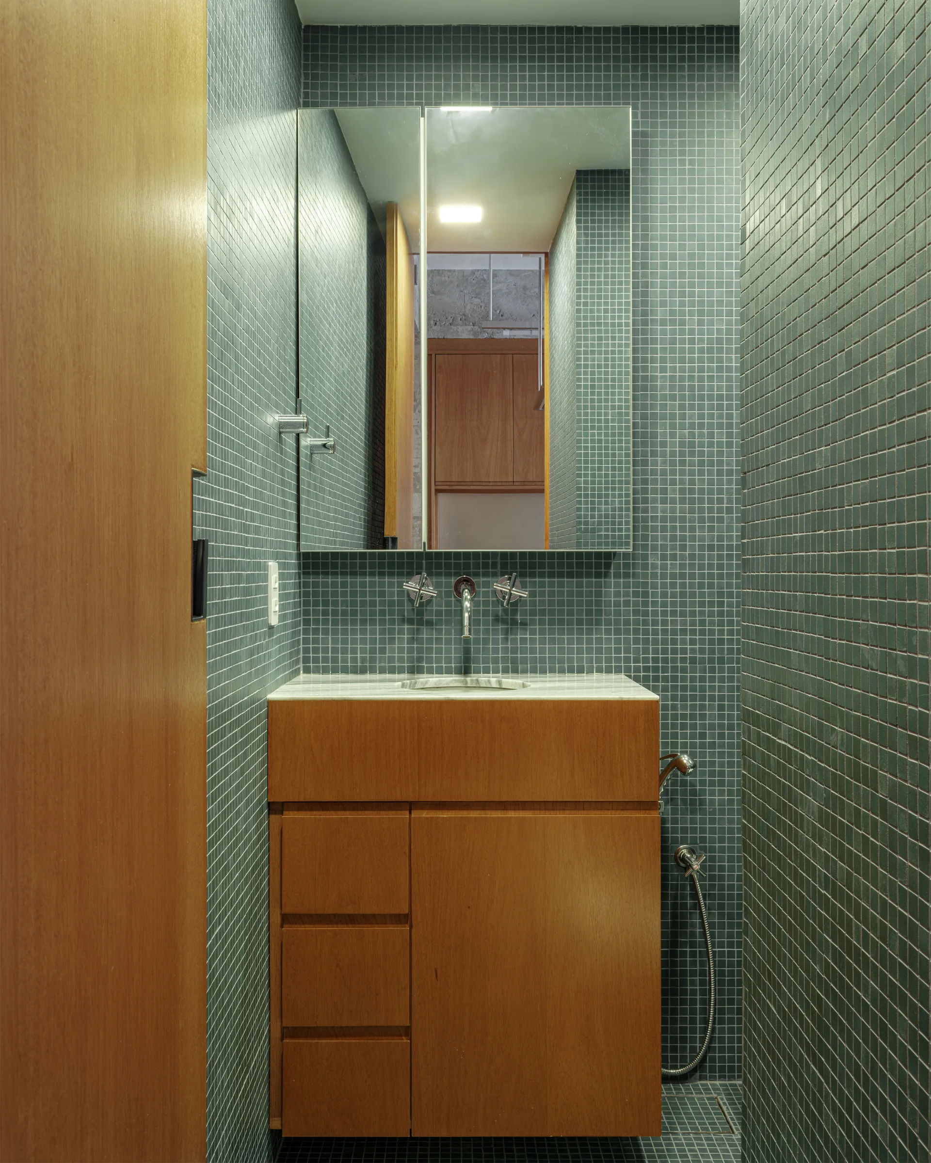 A small bathroom featuring teal square mosaic tiled walls, a wood vanity cabinet with a stone countertop, and a wall-mounted faucet under a mirror.
