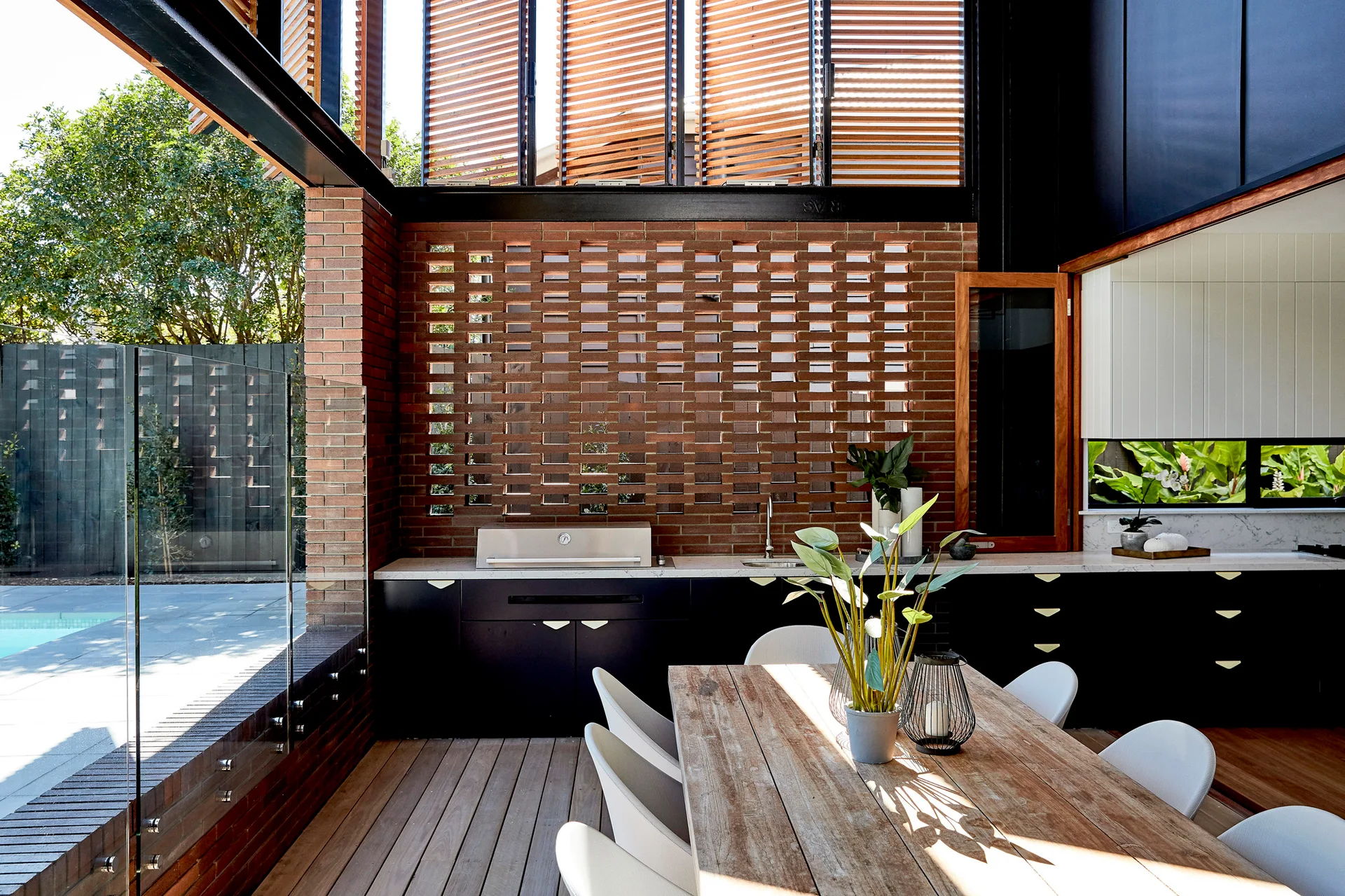 An outdoor dining patio features a weathered wood table with white chairs, black kitchen cabinets, and a perforated red brick screen wall.
