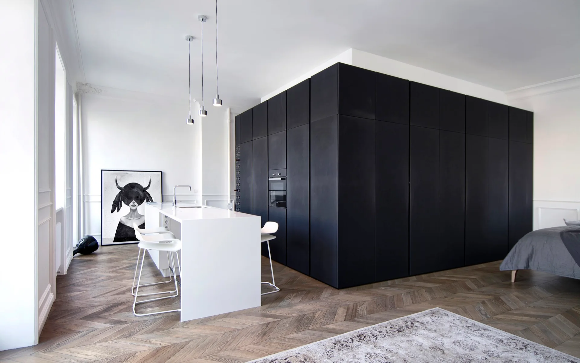 A white kitchen island with stools stands in front of a massive matte black cabinetry block on herringbone wood flooring.