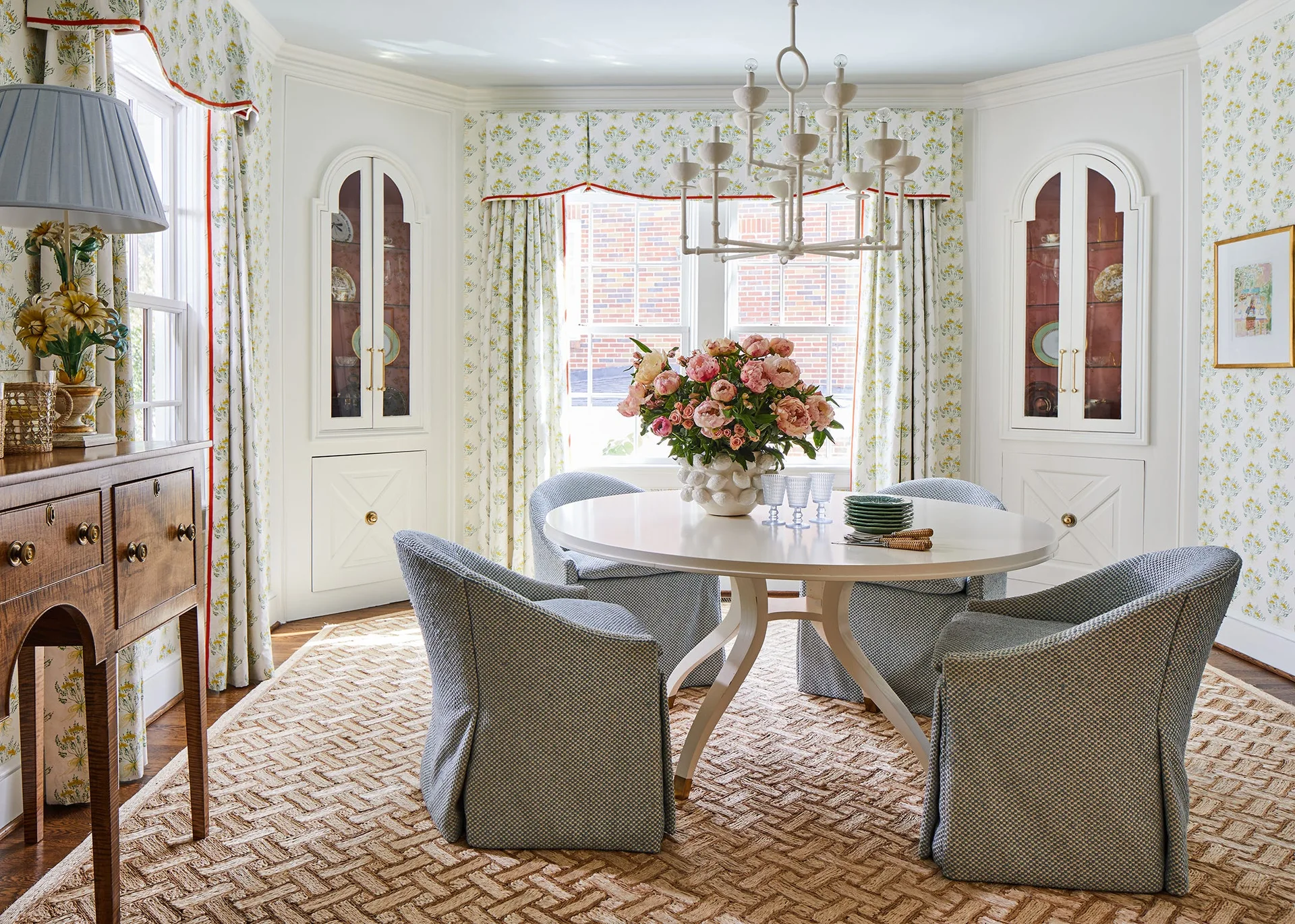 A traditional dining room featuring a round white table, blue upholstered skirted chairs, arched built-in cabinets, and floral wallpaper.