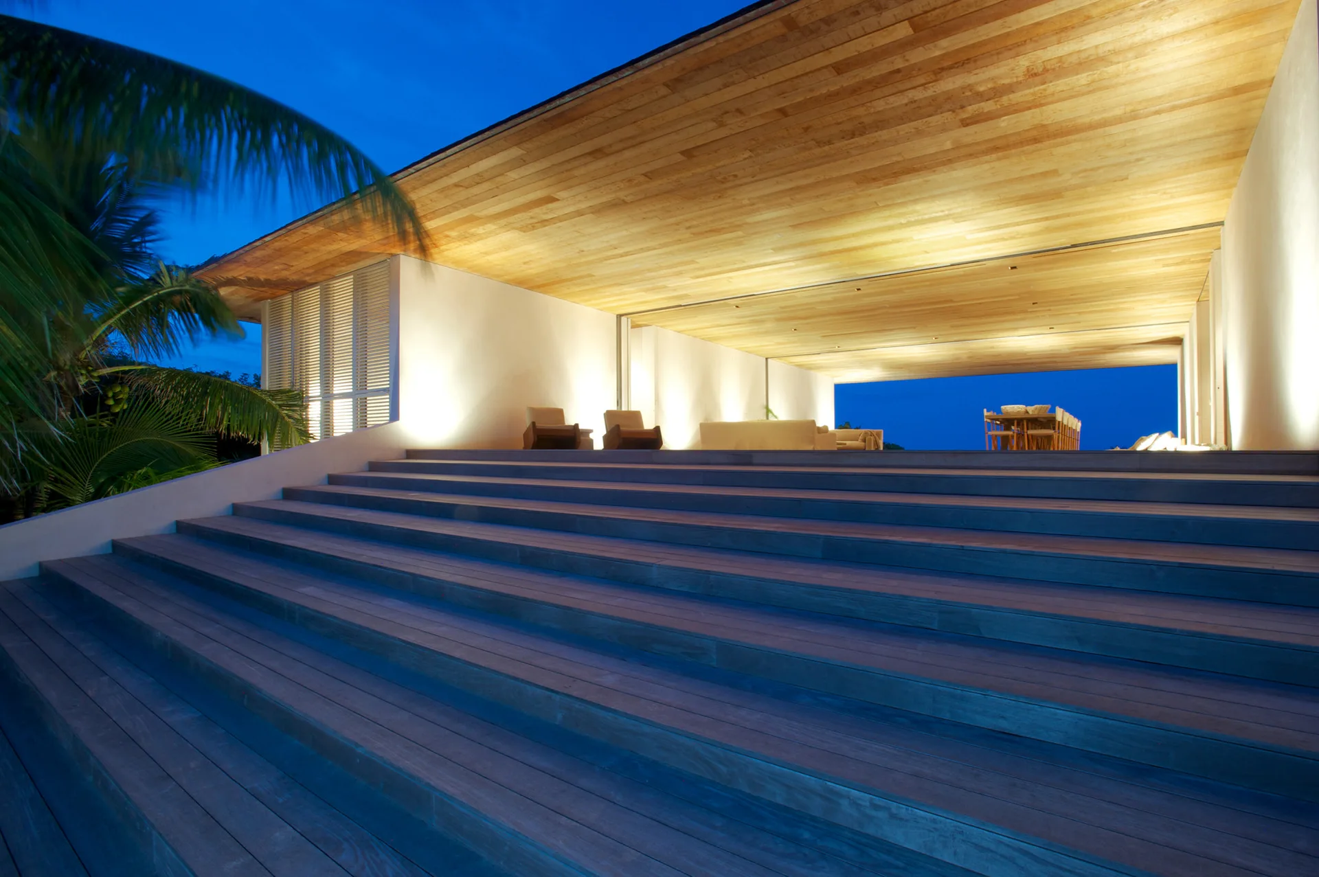 A wide wooden staircase leads up to an open-air modern terrace with white stucco walls, a wood-paneled ceiling, and warm evening lighting.
